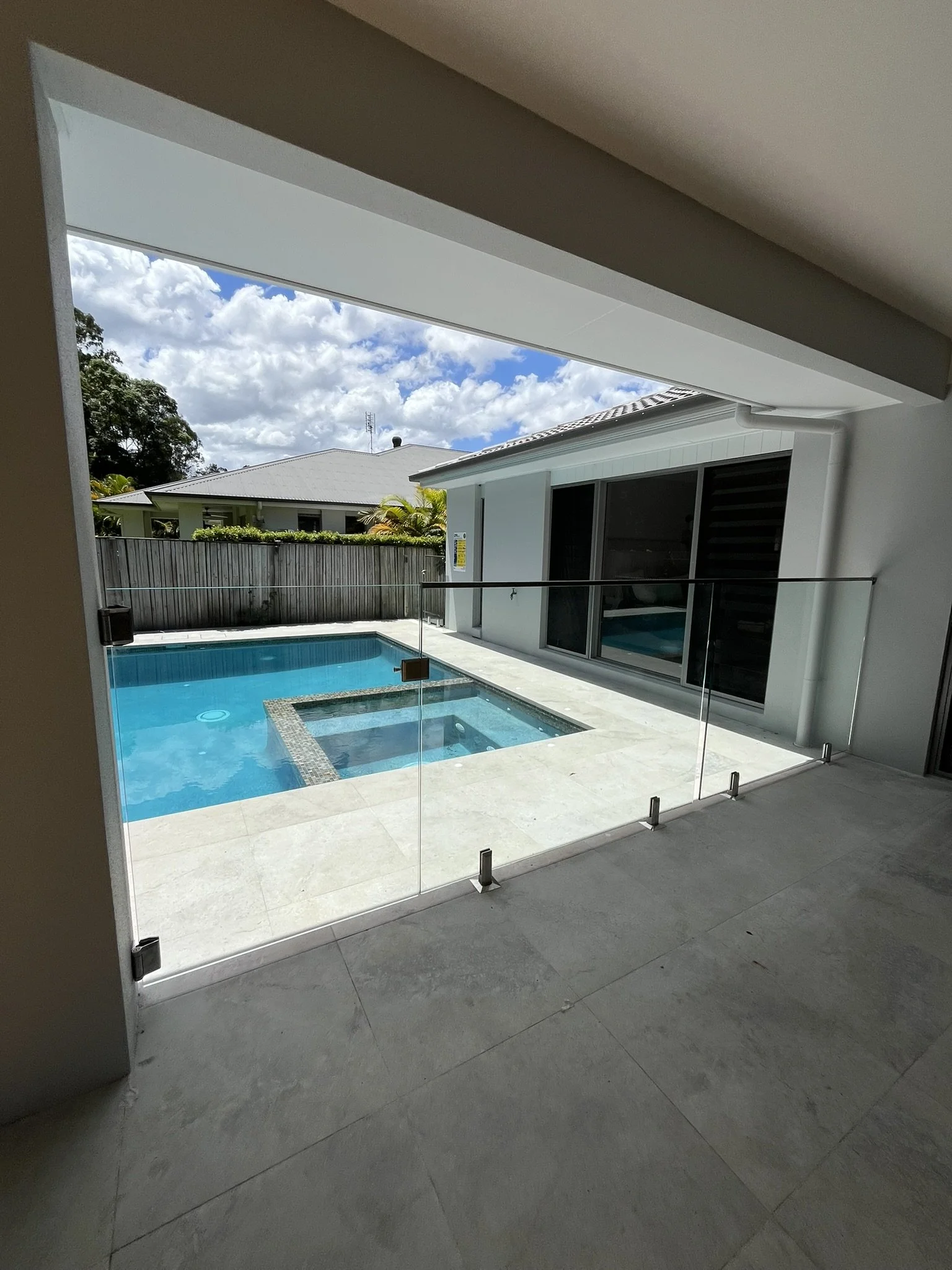 View of a swimming pool and surrounding houses, seen from an open doorway with a glass safety barrier.