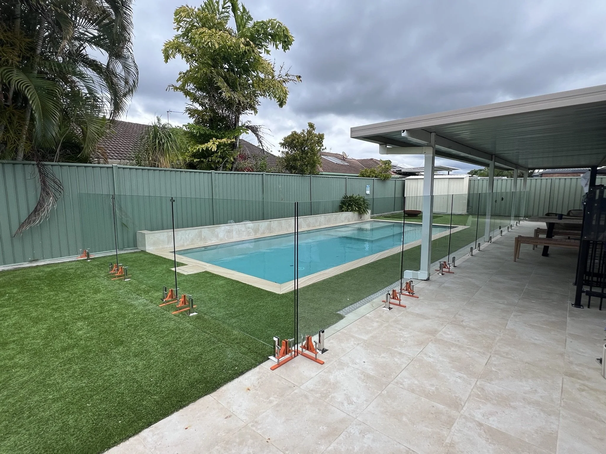 A backyard with a swimming pool enclosed by a glass fence, lush green grass, and tall trees. There is a covered patio area with chairs and a lounge chair, and neighboring houses with rooftops and solar panels are visible under a cloudy sky.