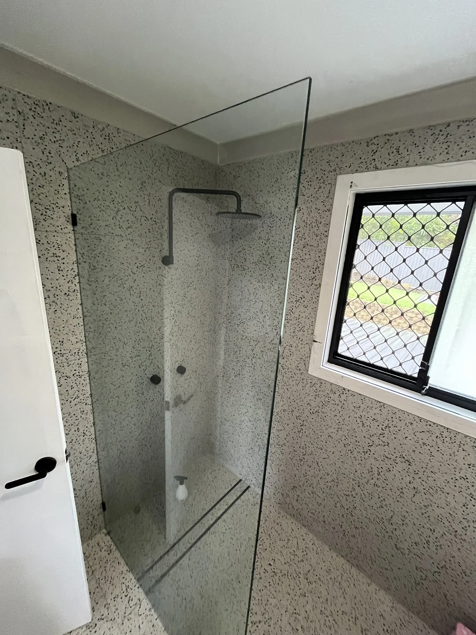 A bathroom with a bed and a window with a security grill, a larges glass shower enclosure, and a terrazzo floor.