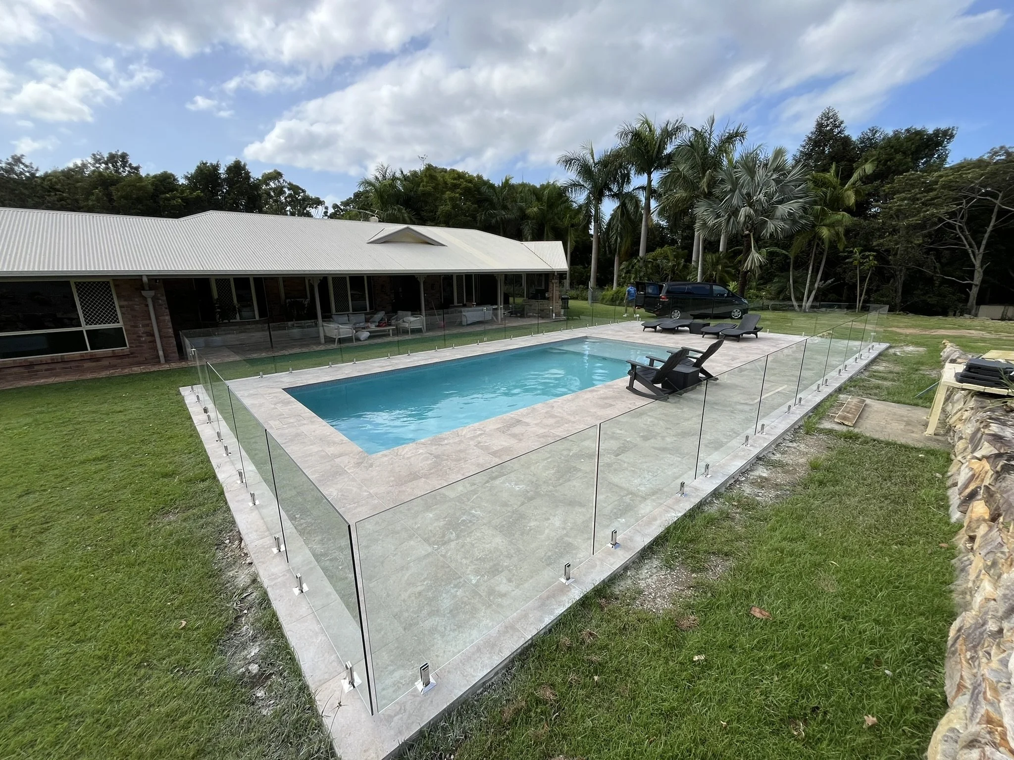 A backyard with a swimming pool surrounded by a glass fence, lounge chairs, and a house with a white metal roof and a covered porch, with trees and a cloudy sky in the background.