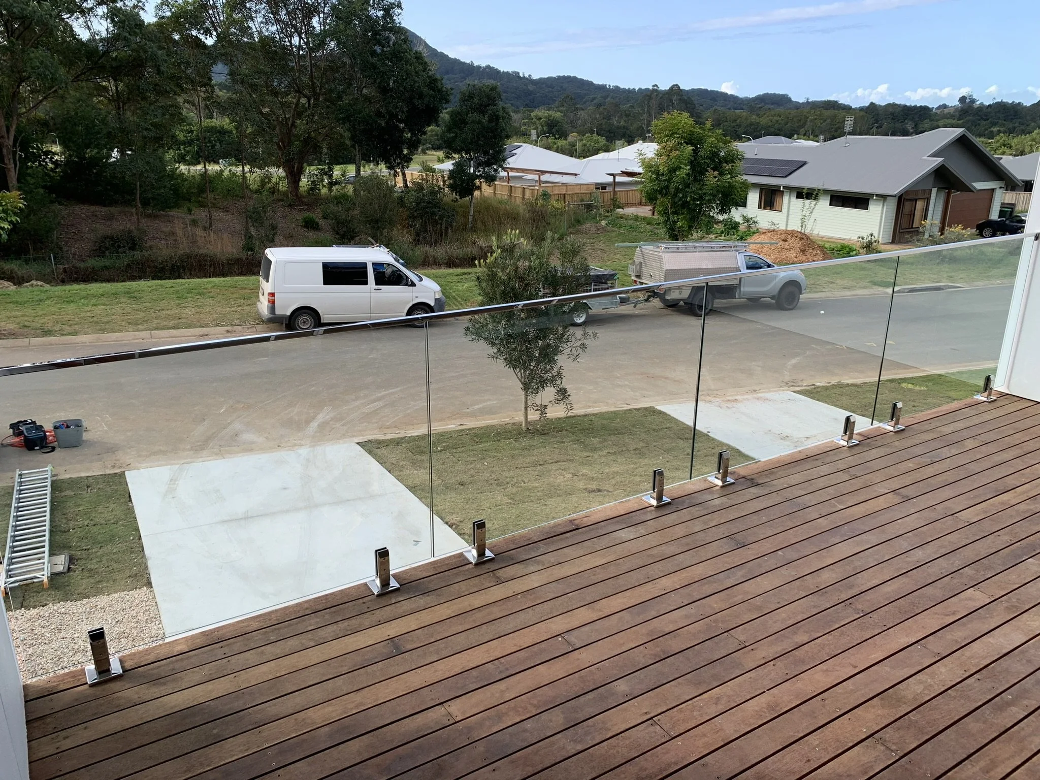View from a balcony with a glass railing, looking out over a suburban neighborhood with houses, trees, a parked white van and a truck, and hills in the background.