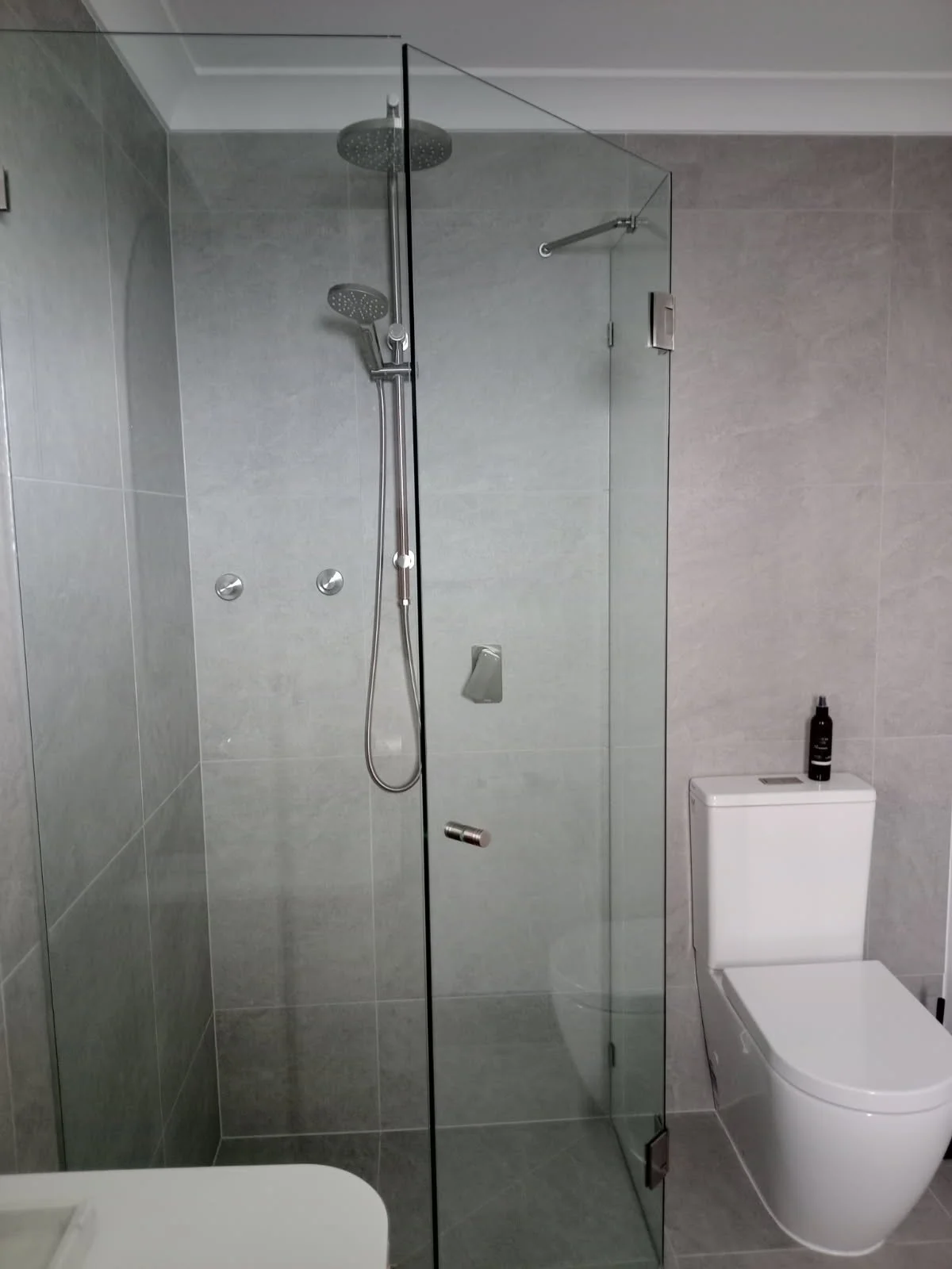 Modern bathroom with a glass shower enclosure, rain shower head, handheld shower, and a white toilet with a black bottle on top.