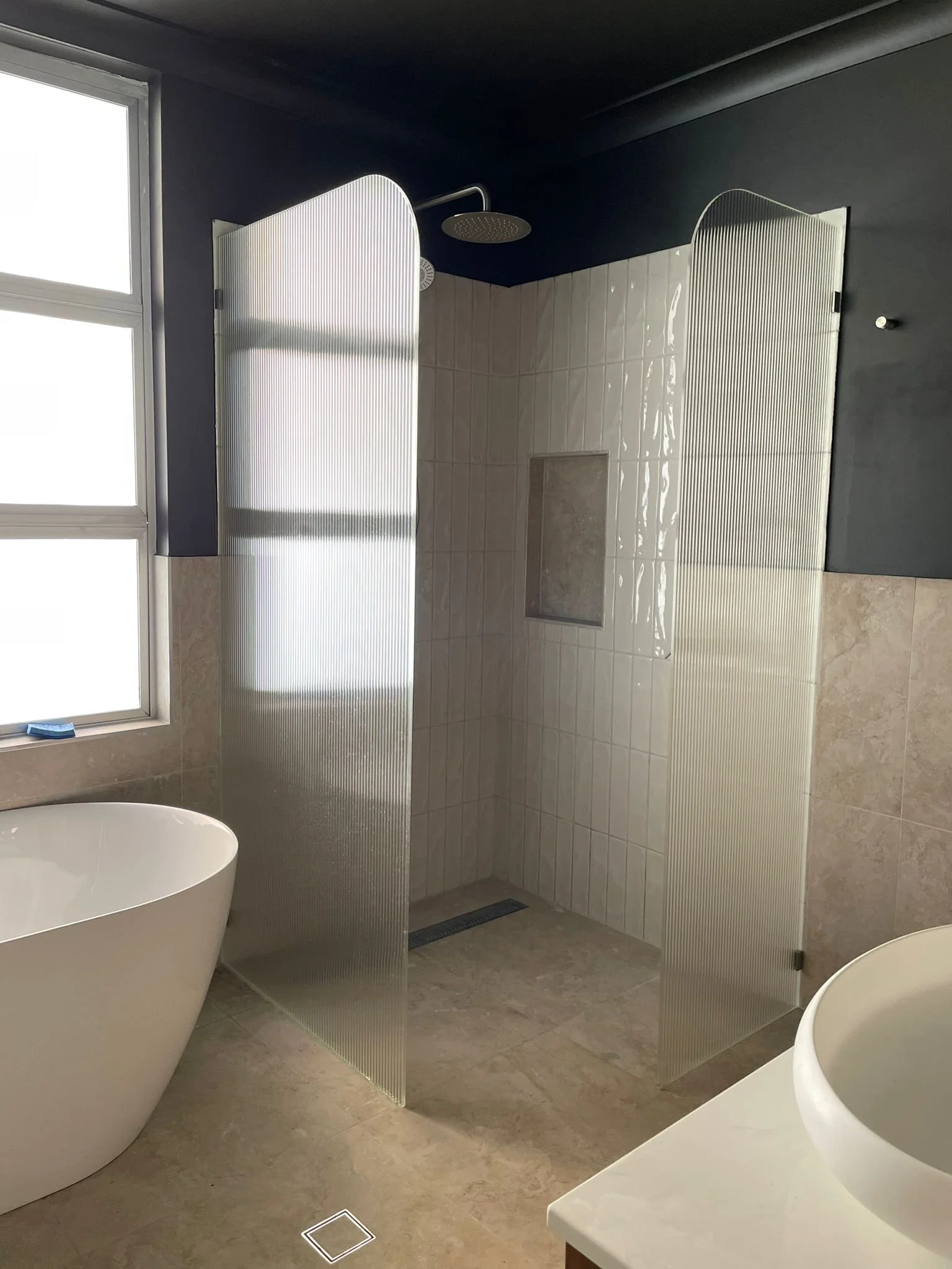 Modern bathroom with a freestanding bathtub, a walk-in shower with two frosted glass partitions, beige tiled walls, and a window letting in natural light.