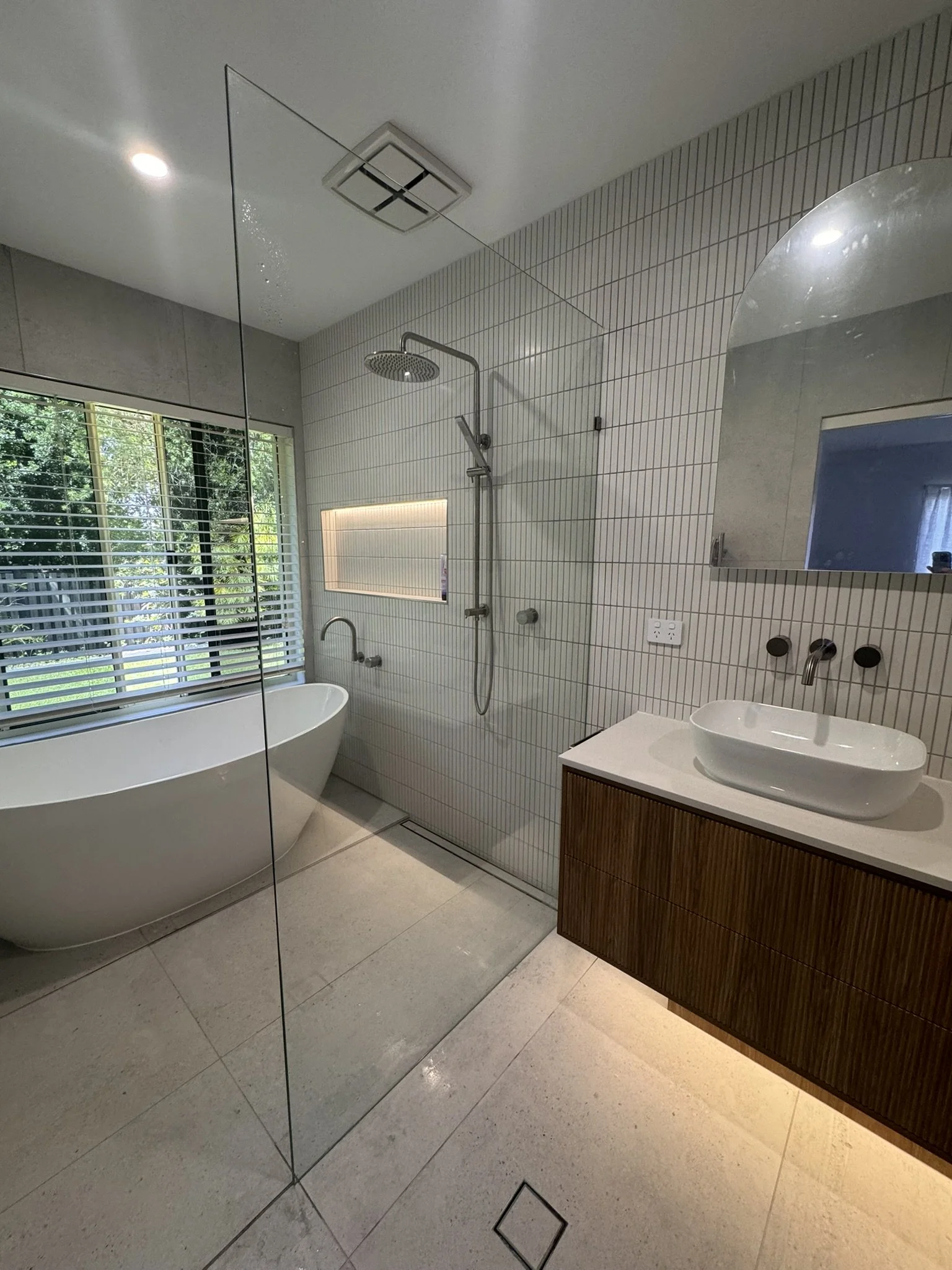 Modern bathroom with freestanding bathtub, walk-in shower with rain shower head, large mirror, and window with blinds.