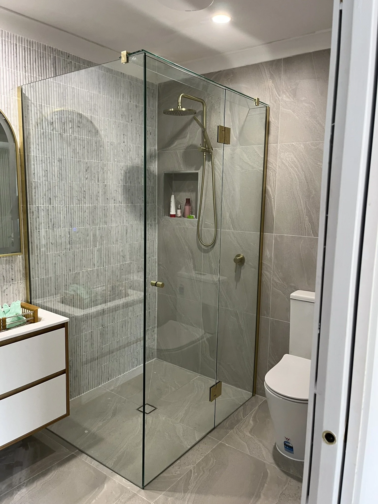 Modern bathroom with glass shower enclosure, gray marble walls, a white toilet, and a wooden vanity with organized toiletries. Recessed ceiling light illuminates the space.