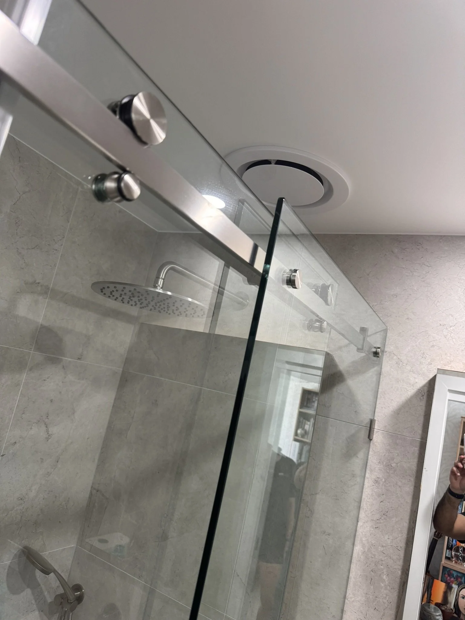 A view of a bathroom with a shower, glass door, and a beige tiled wall. The showerhead and control knobs are visible.