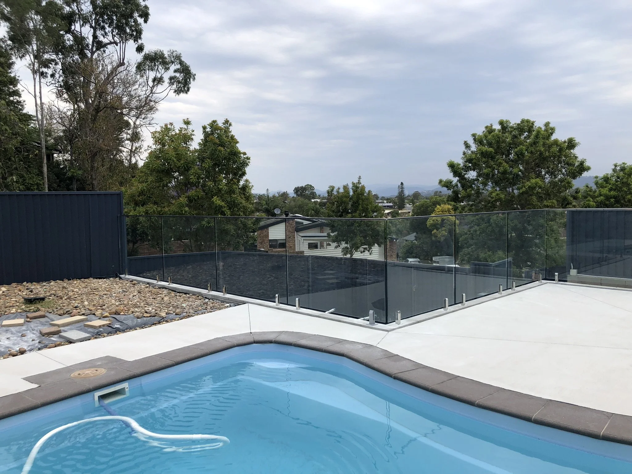 A backyard with a swimming pool, a fenced patio, and a view of trees and houses under a cloudy sky.