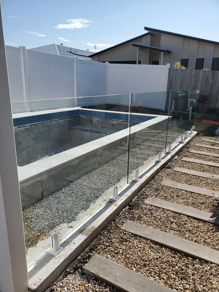 Under-construction swimming pool with glass fencing, surrounded by a gravel and wooden pathway, in a backyard with a house and privacy fence in the background.