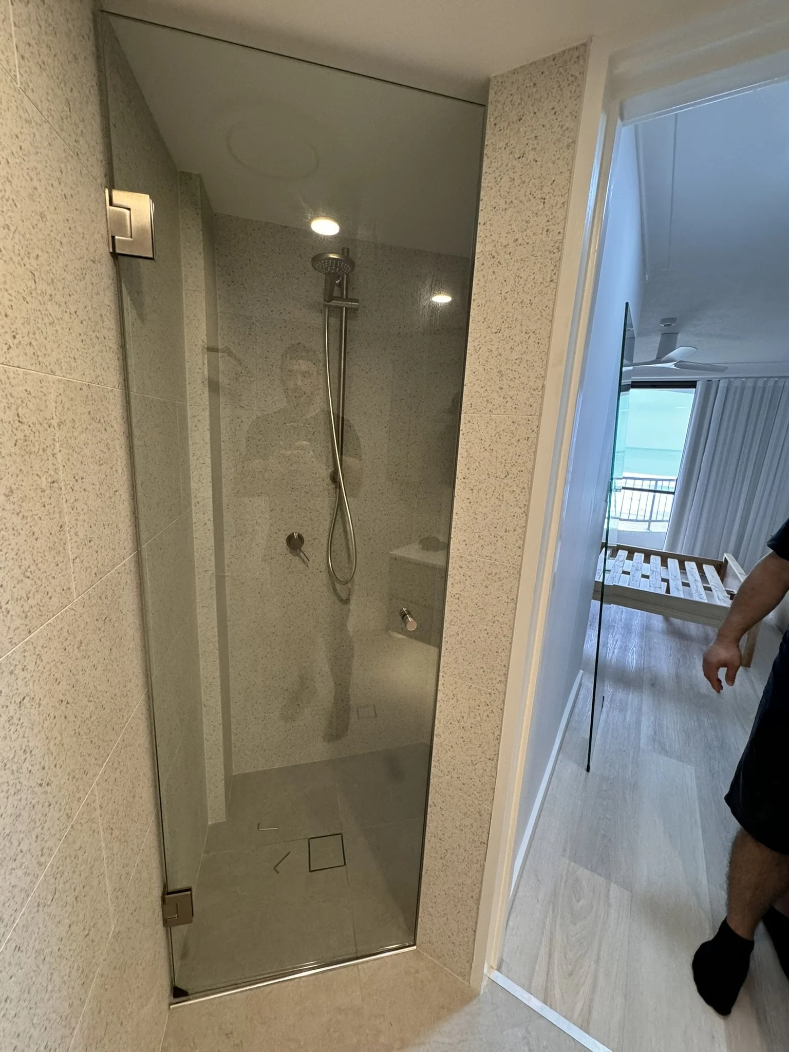 A modern bathroom shower with a glass door, a rainfall showerhead, and beige tile walls, seen from a reflection in a mirror.