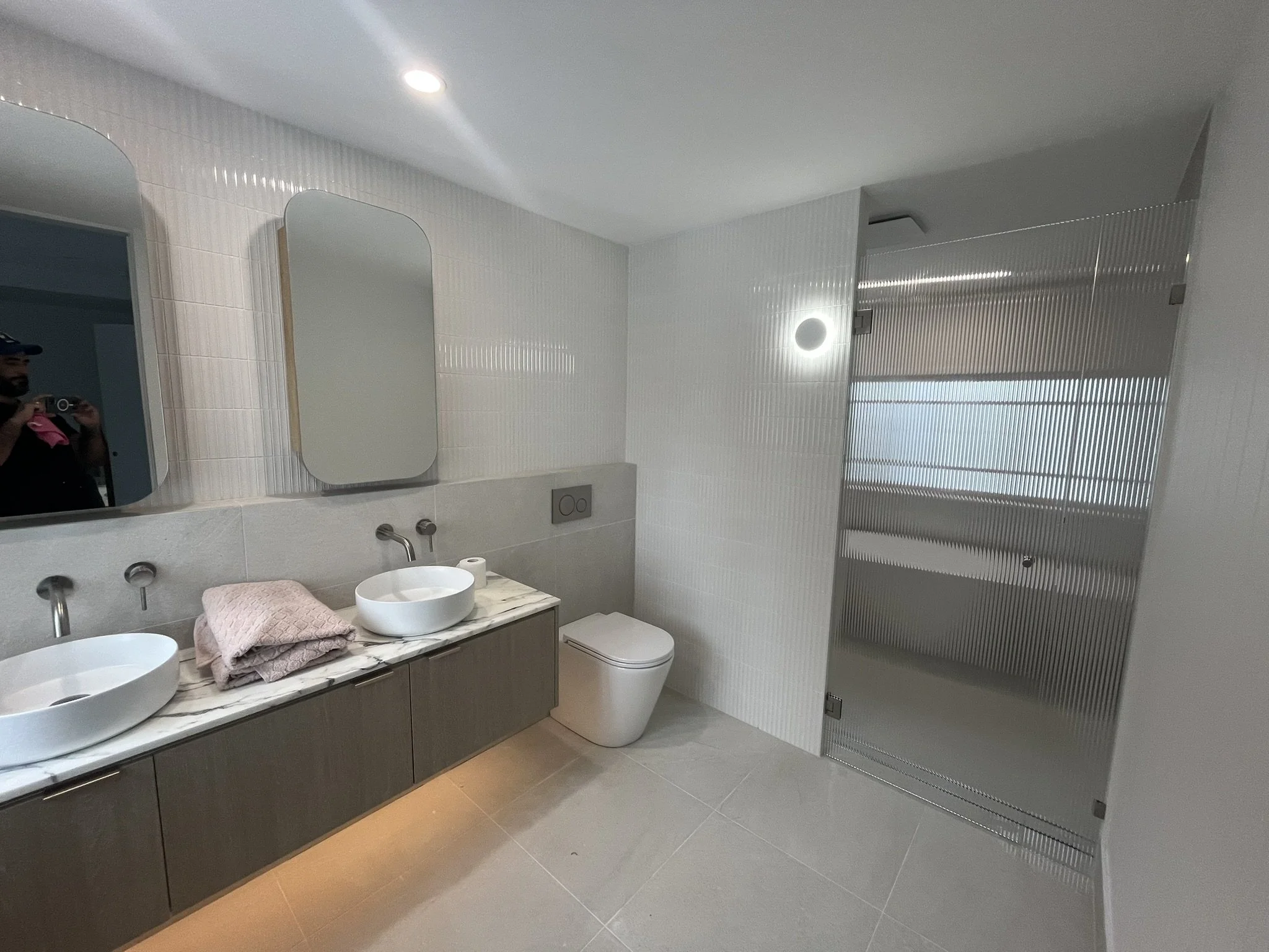 Modern bathroom with double sinks on marble countertop, mirror, white wall tiles, toilet, and a glass shower enclosure with frosted glass panels.