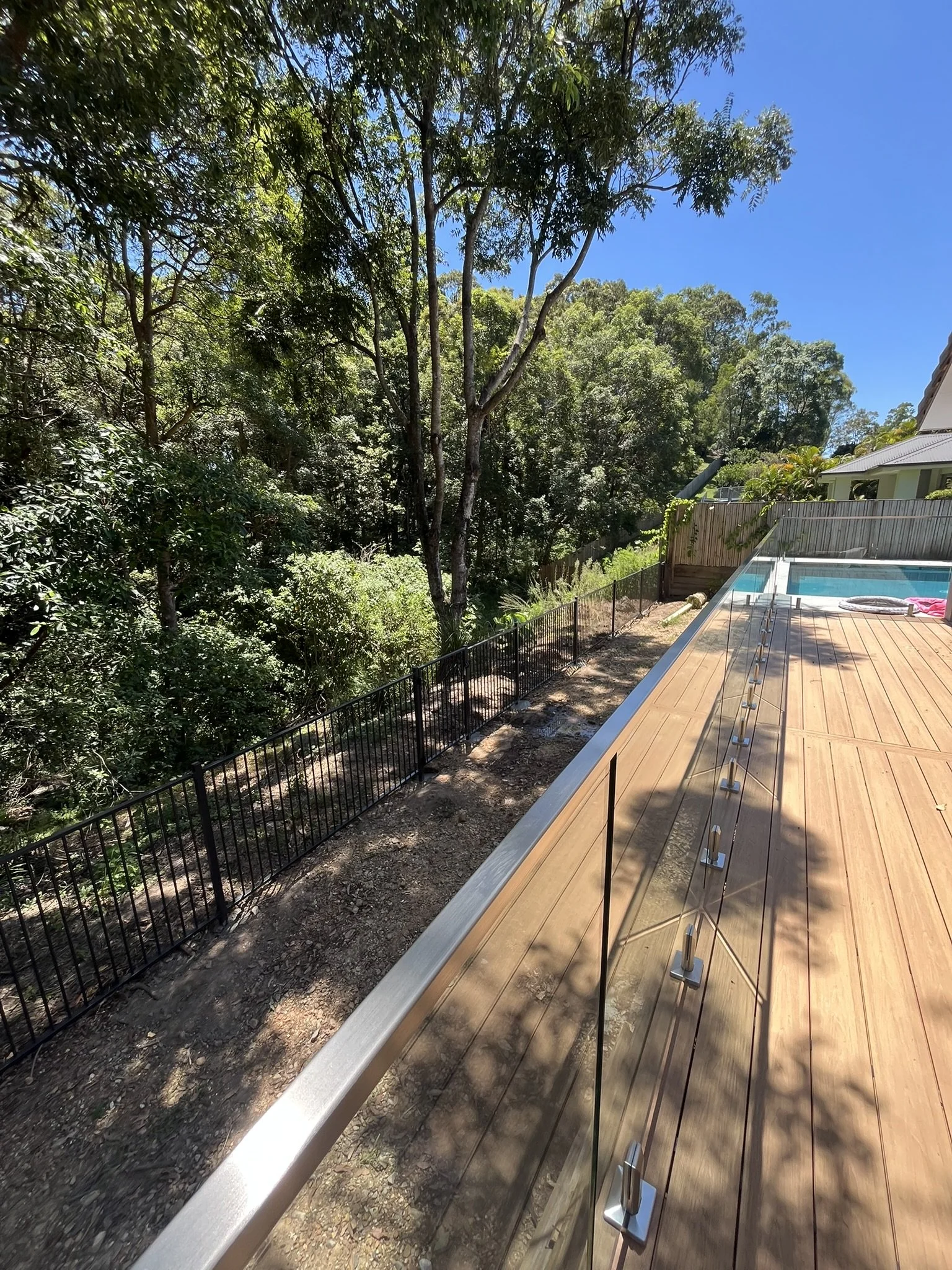 A wooden deck with glass railing next to a backyard pool, overlooking a lush green wooded area with trees and a blue sky.
