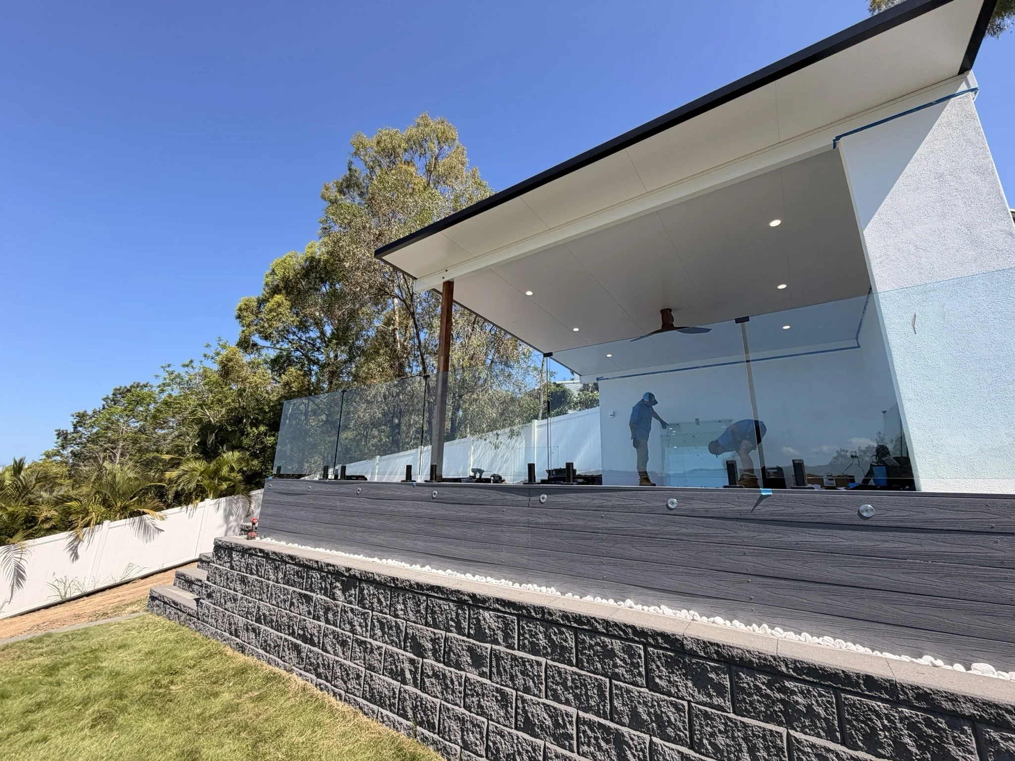 Modern house with an overhanging roof and glass balcony, two people working near the house, surrounded by trees and a bright blue sky.