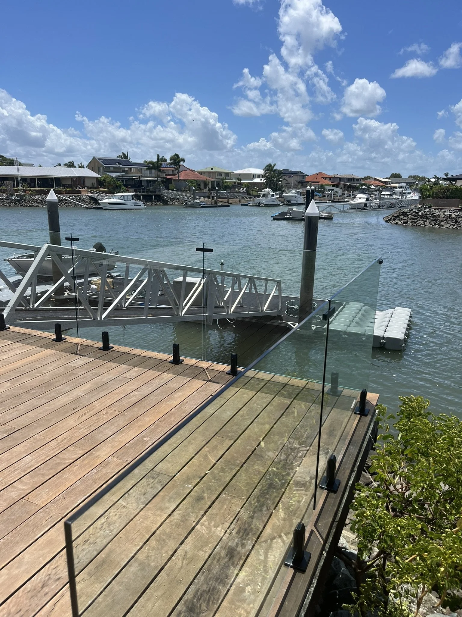 A waterfront view of a small marina with several boats docked along a wooden pier. Residential houses with colorful roofs line the opposite shore under a bright blue sky with scattered white clouds.