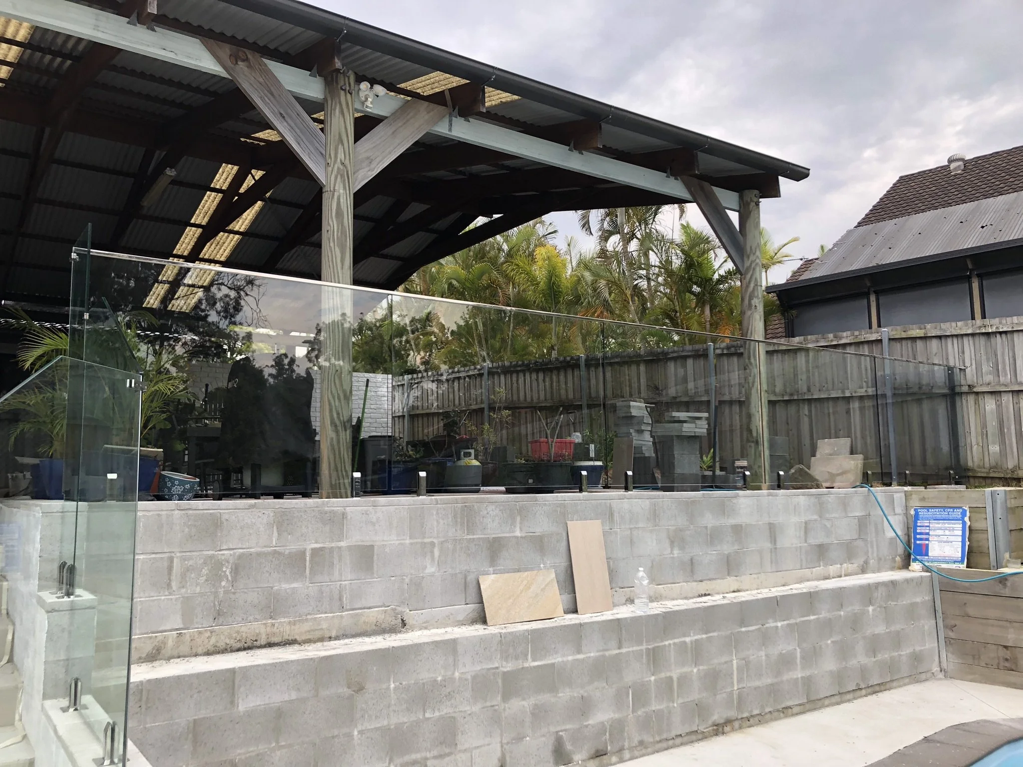 Construction site with a concrete block wall, glass panels, and a wooden structure with metal roofing, near a house with a tiled roof and trees in the background.
