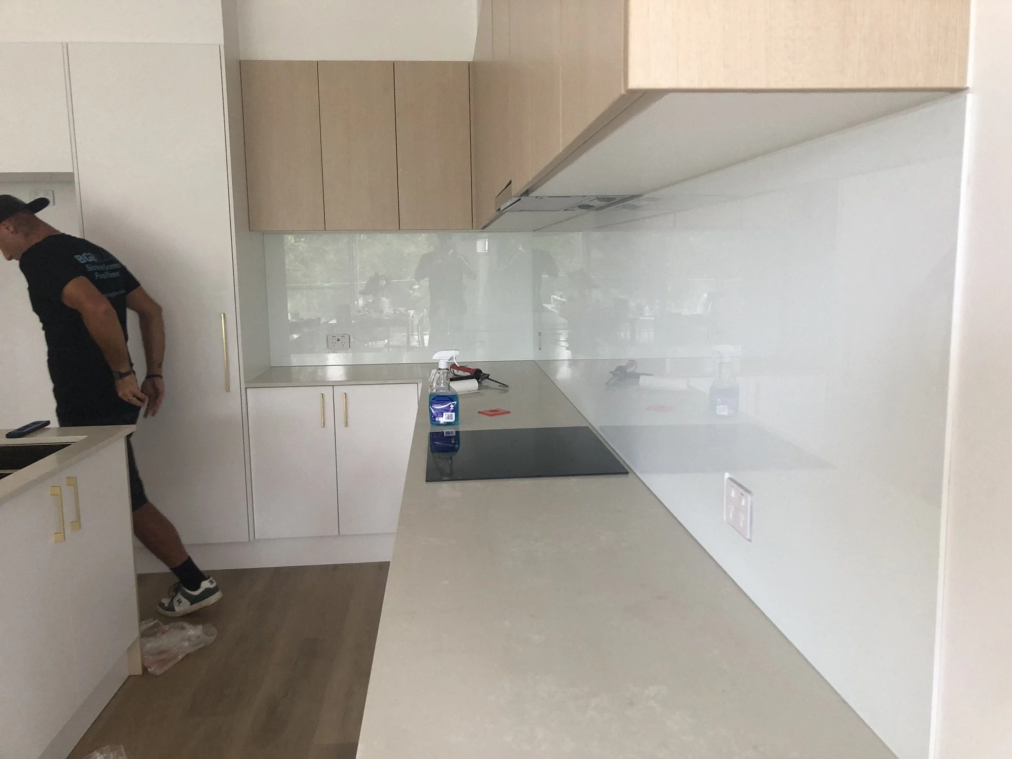 A modern kitchen with white and light wood cabinetry, white countertops, and a reflective backsplash. Two people are in the kitchen; one person is standing near the left side wearing a black t-shirt, black shorts, and sneakers, with part of their fac