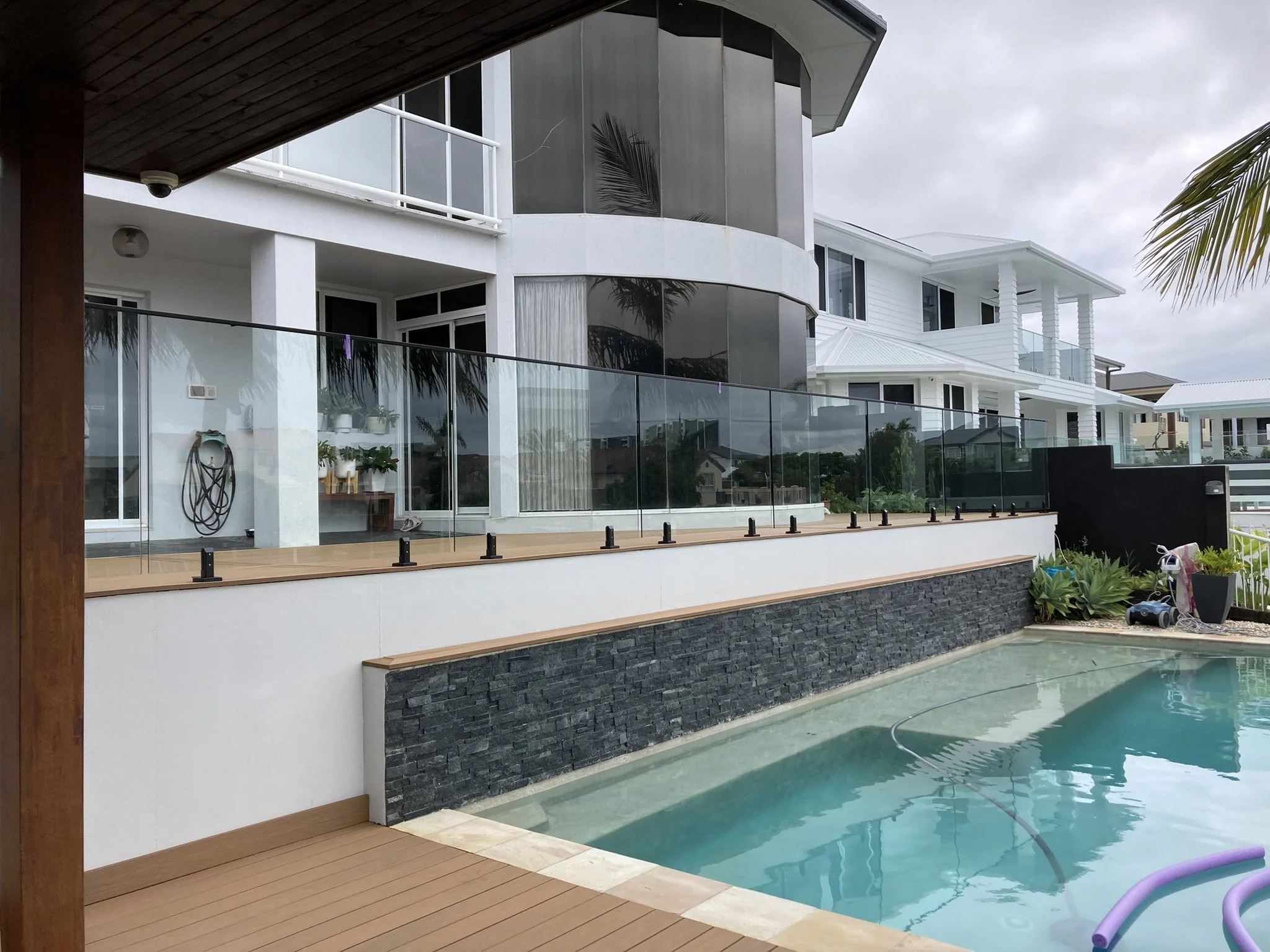 Modern house with white exterior, large glass windows and balconies, overlooking a pool with wooden decking and a brick wall.