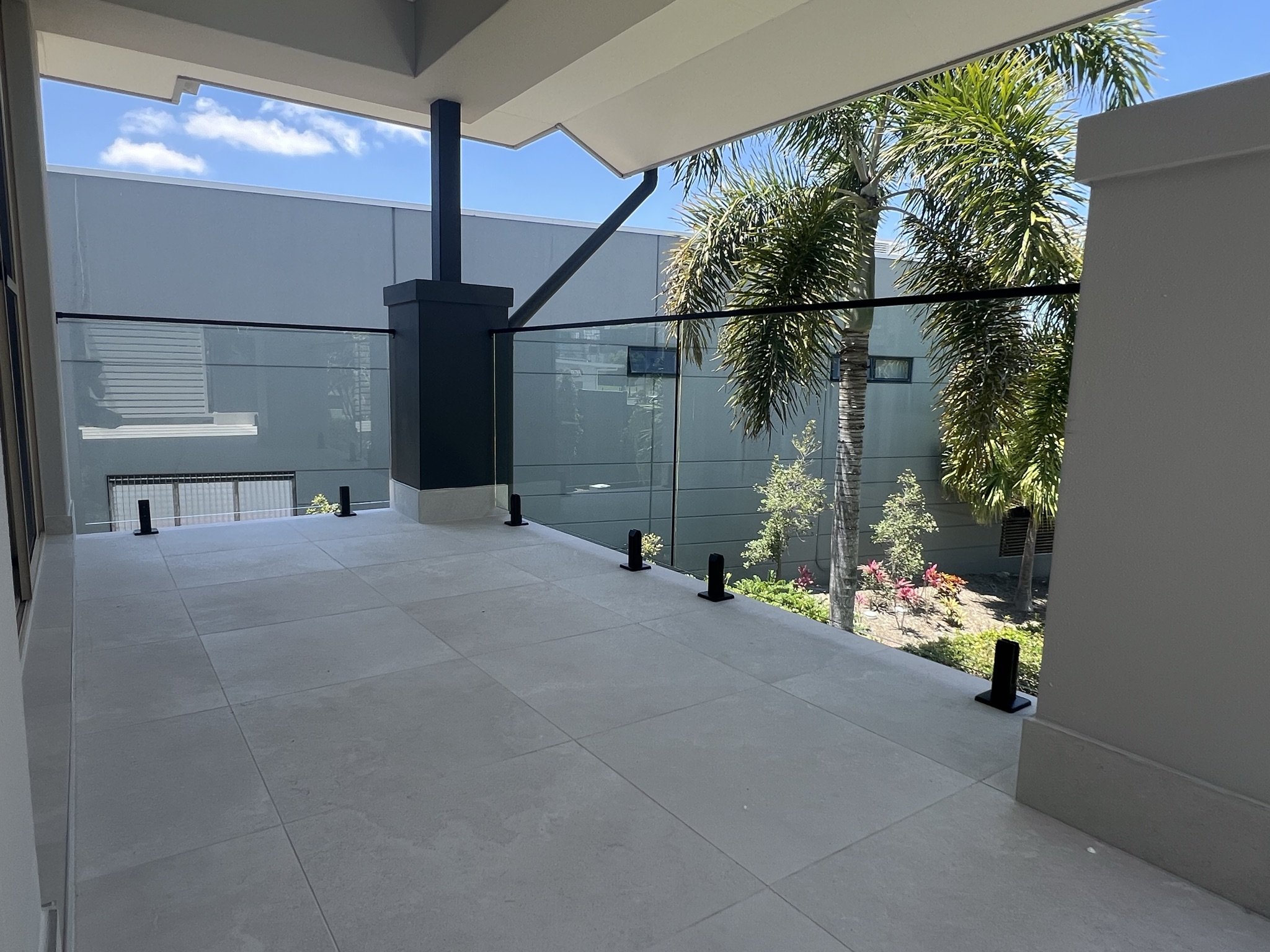 View from a balcony with glass railing, grey tiled floor, white ceiling, palm trees outside, blue sky with a few clouds.