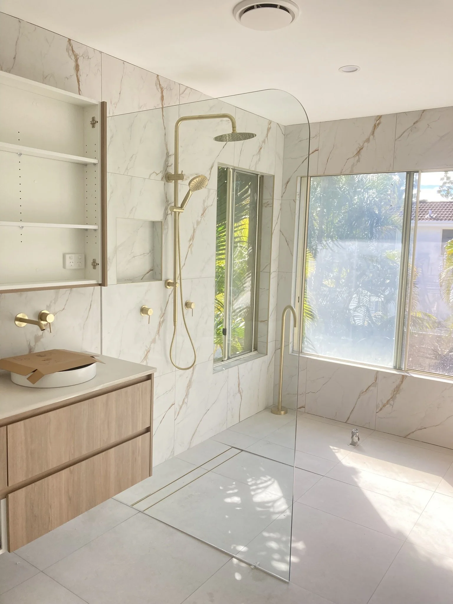 Modern bathroom with marble walls and floor, a glass shower enclosure, brass fixtures, a window with a view of greenery, and a minimalist wooden vanity.
