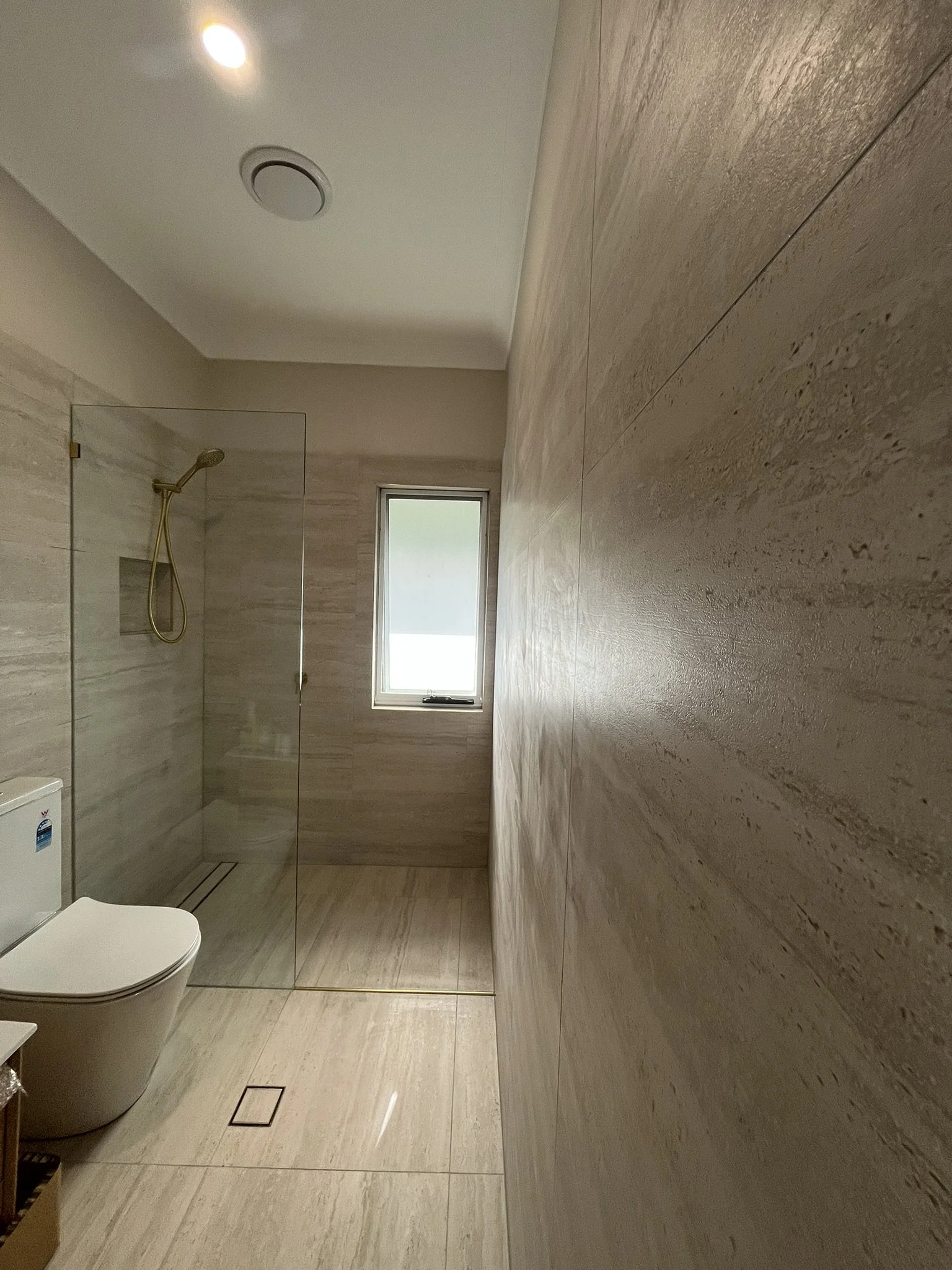 A modern bathroom with a toilet, a walk-in shower with glass panel, a window, and beige tiled walls and floor.