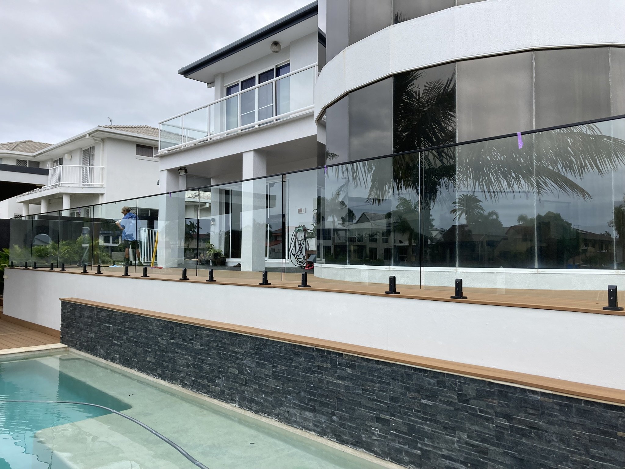 Modern multi-story house with glass balcony railings and a white exterior, adjacent to a swimming pool with a black stone wall. A man in a blue shirt and hat is working on the outdoor area.