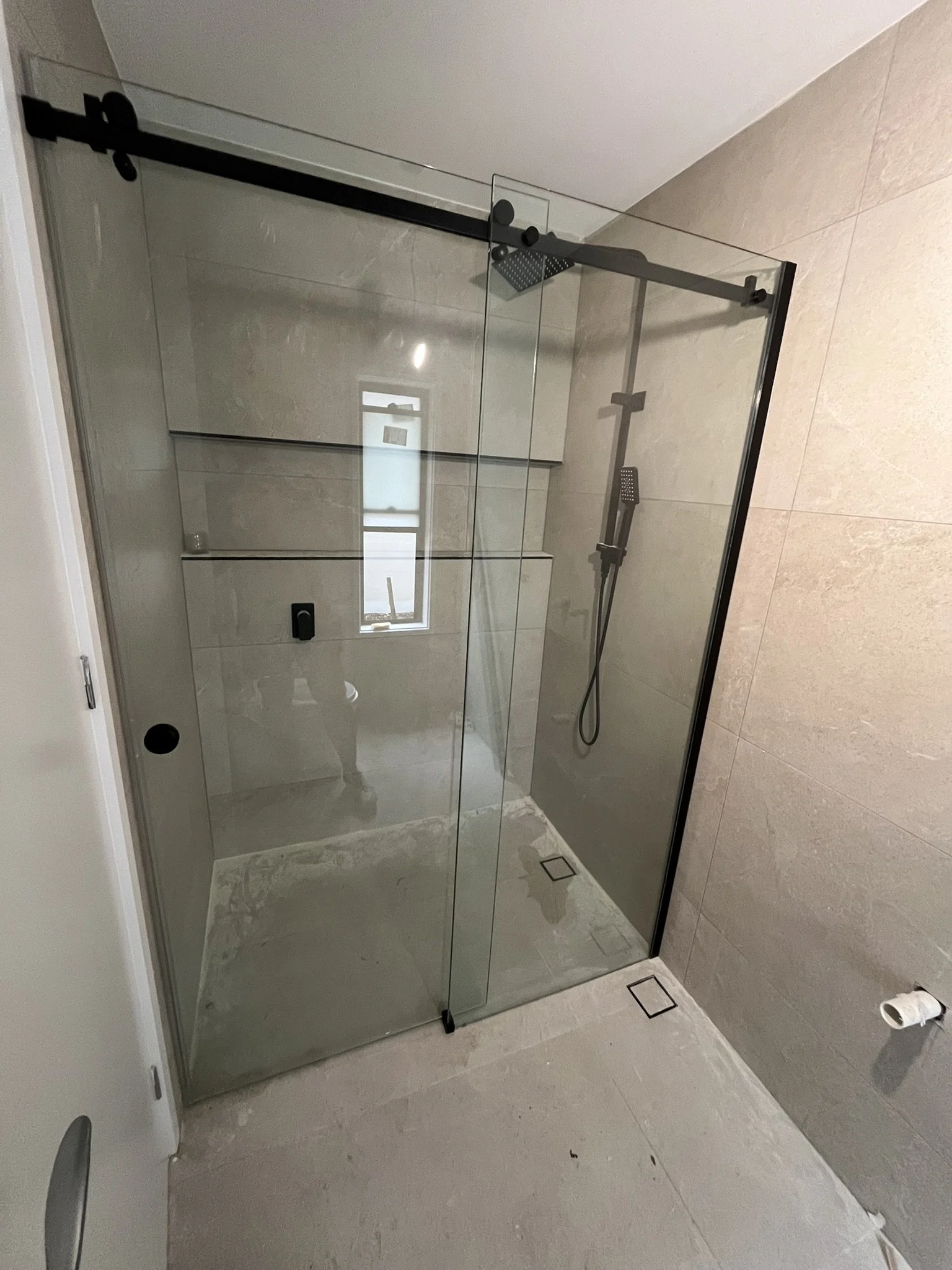 A modern indoor bathroom shower area with a glass enclosure, showerhead, and a small window, with beige tiled walls and floor.