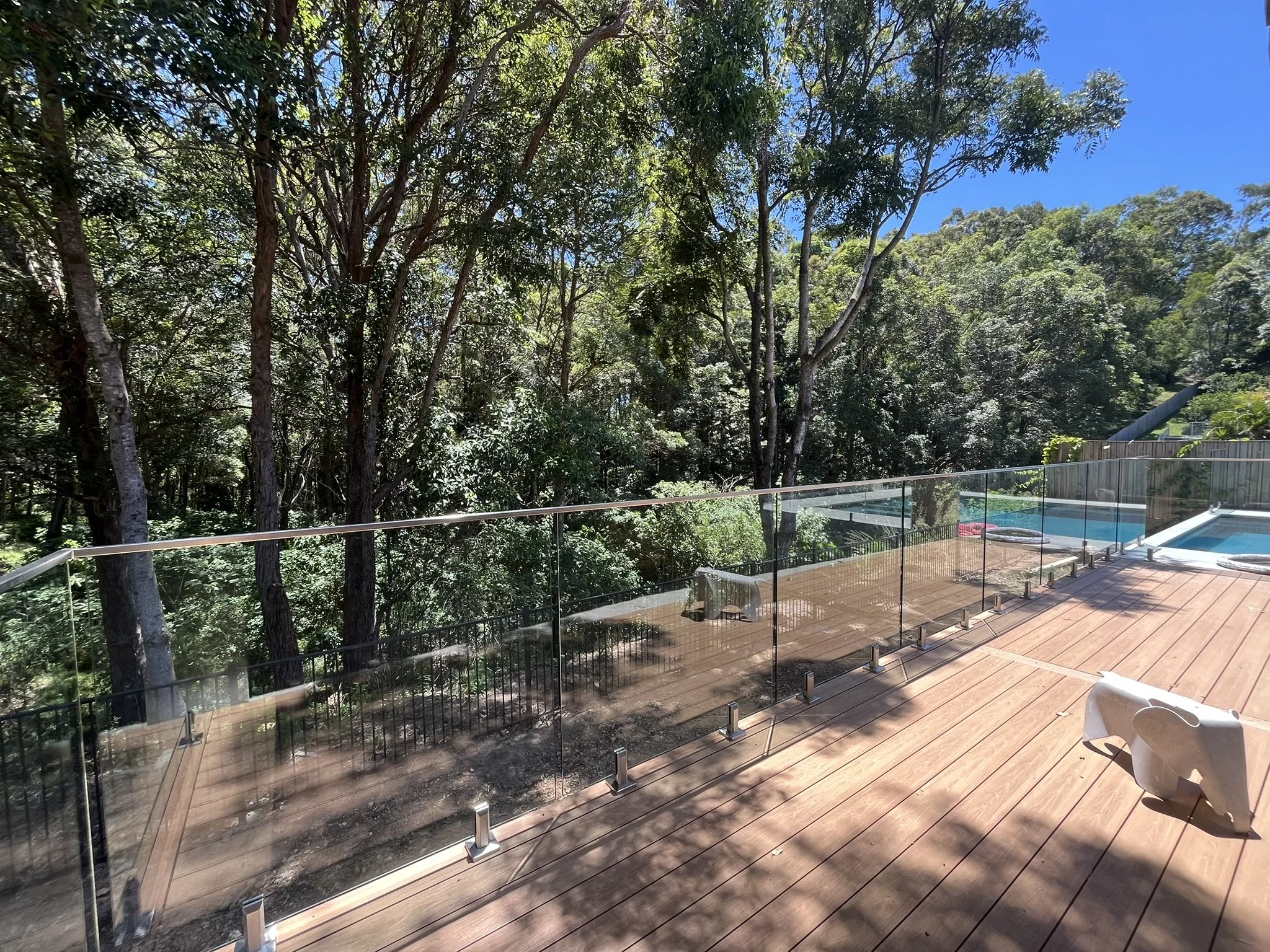 View from a terrace with glass railing overlooking a backyard with trees and a swimming pool, under a bright blue sky.