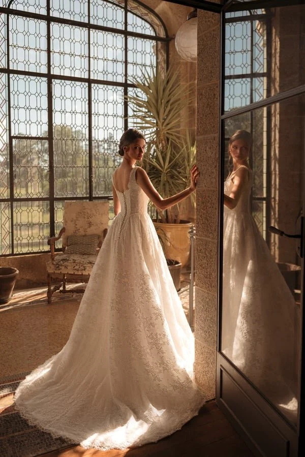 A woman in an elegant white wedding dress standing indoors near a tall window and a mirror, with sunlight streaming in, and a potted plant nearby.