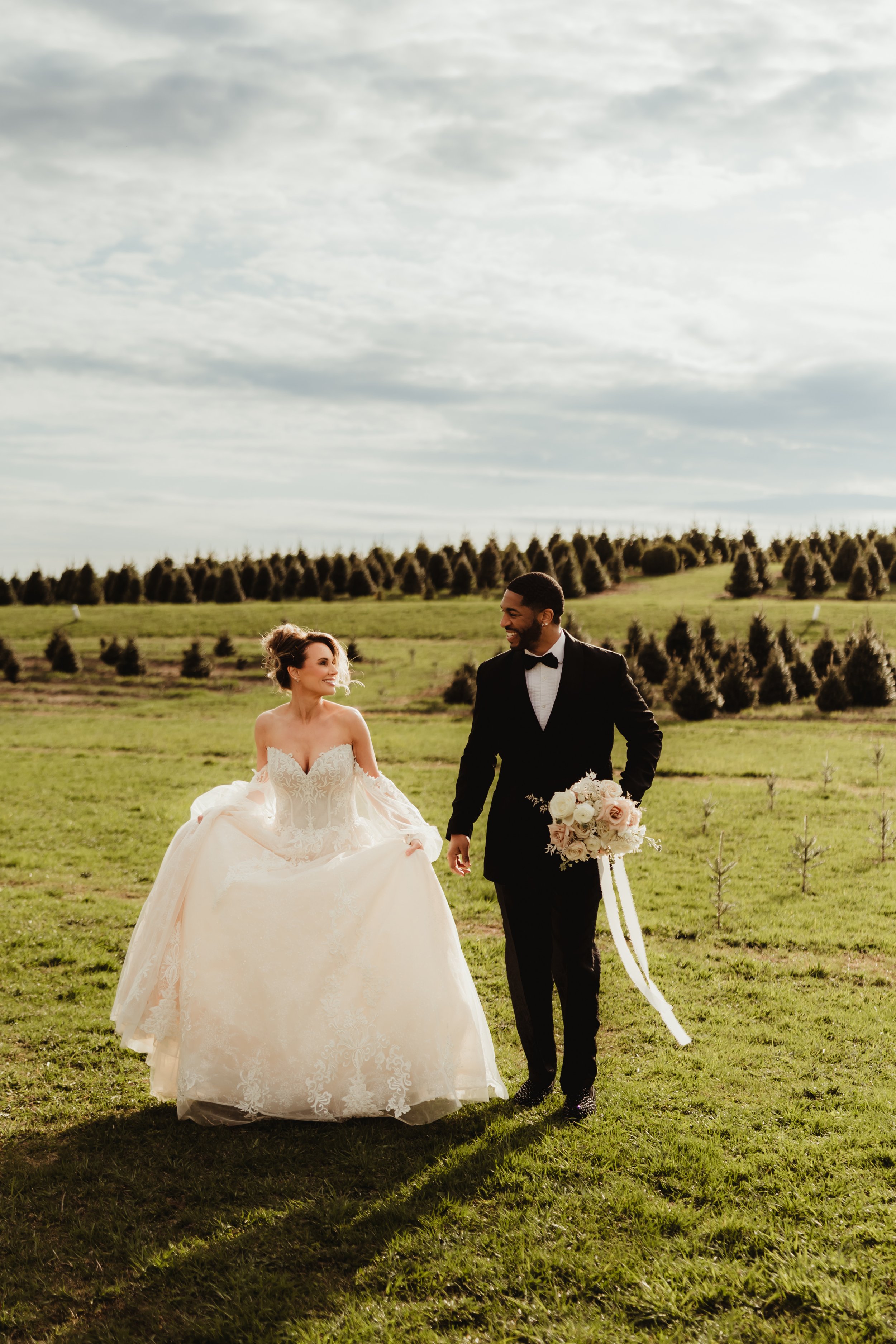 A bride and groom walking together outdoors in a grassy field, with trees in the background and cloudy sky above, during their wedding day.