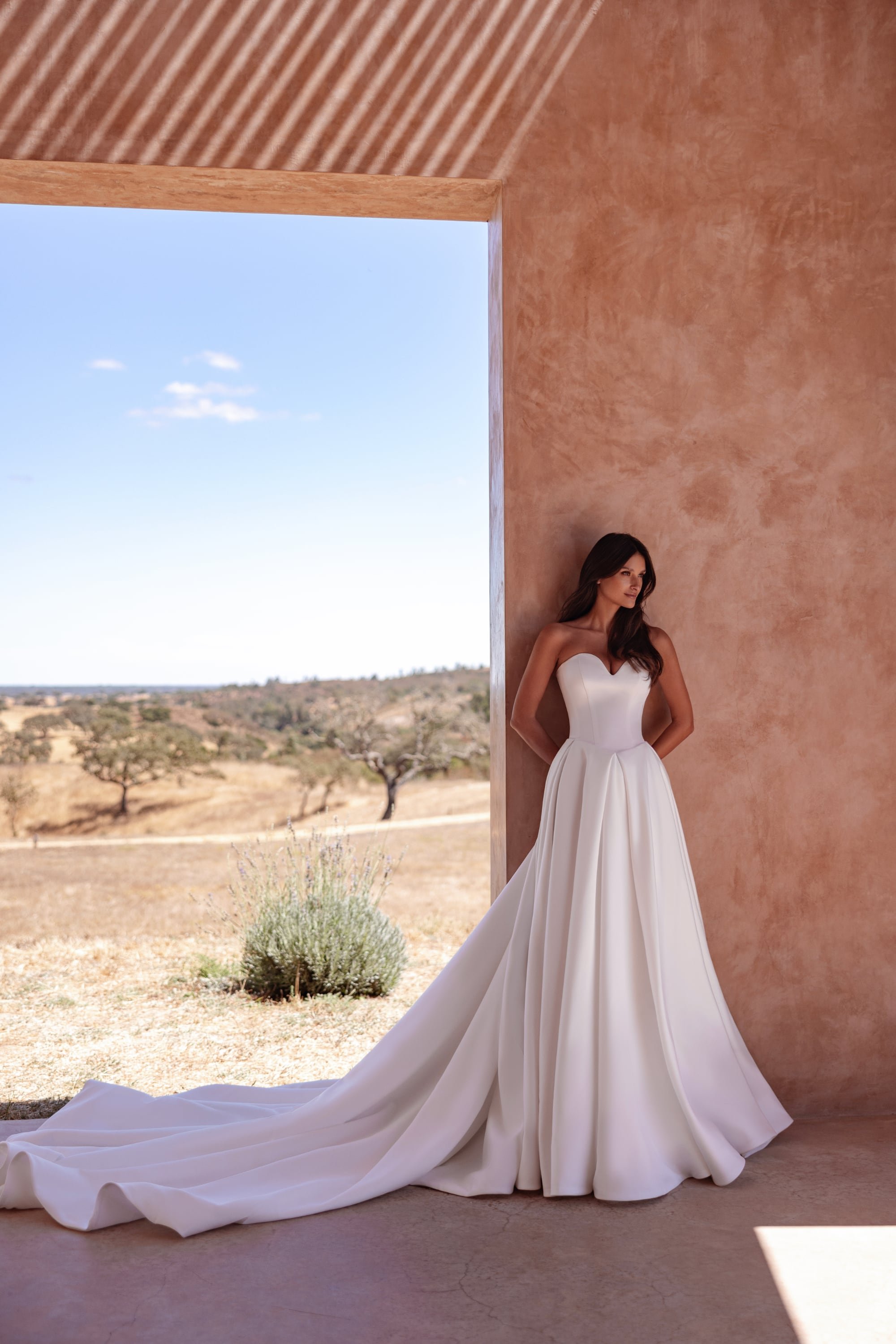 A woman in a strapless white wedding dress stands against a textured coral-colored wall, with a large train flowing onto the ground. She looks to the right, with a landscape of trees and open land visible through an opening in the wall.