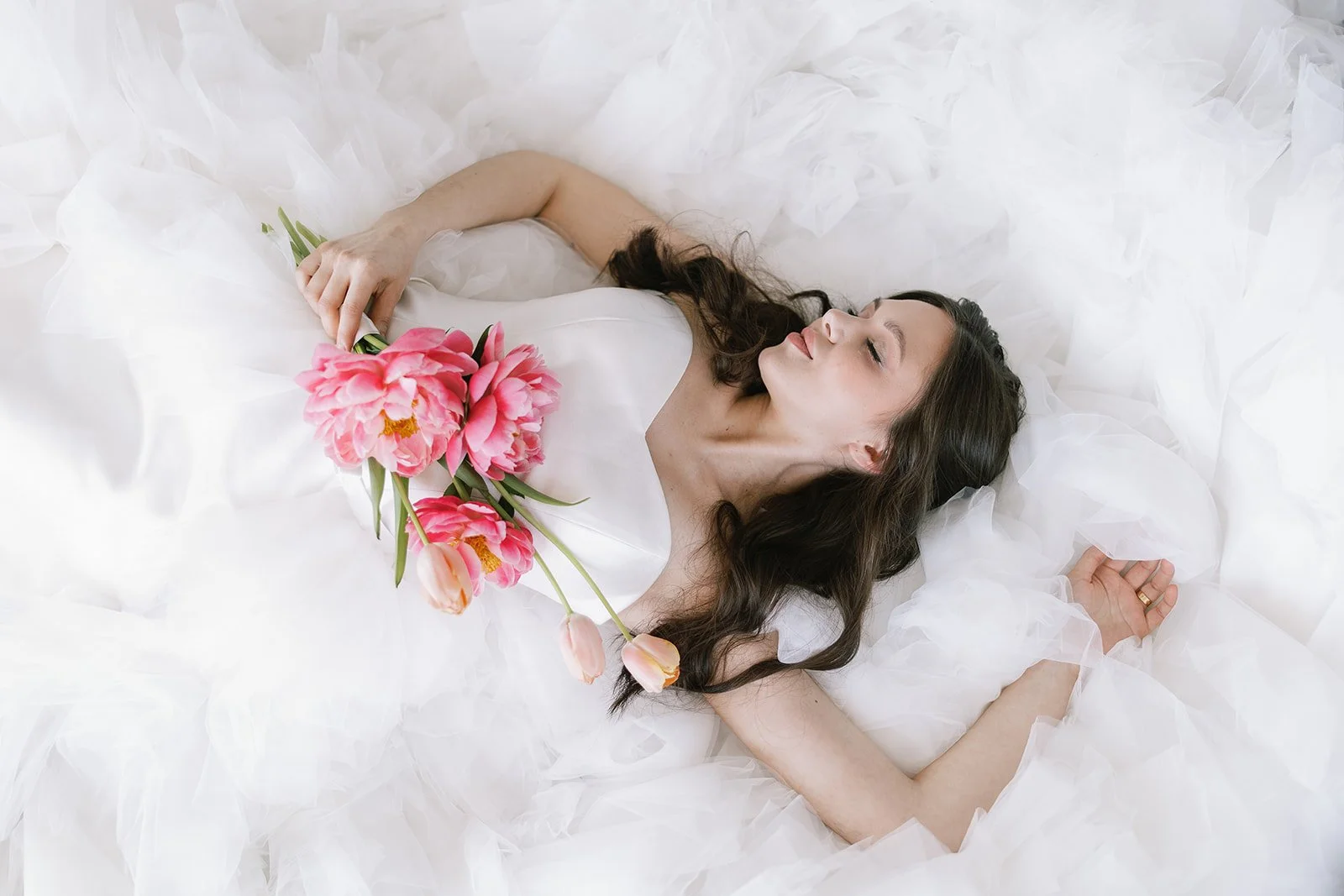 A woman lying on a bed in a white dress, holding a bouquet of pink and peach tulips, surrounded by white tissue paper.