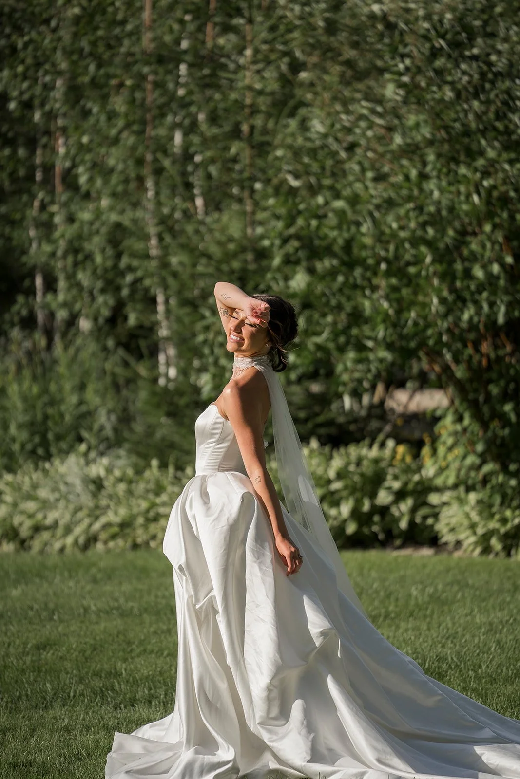 A woman in a white wedding dress standing outdoors on grass with a backdrop of green trees, smiling and shielding her eyes from the sun with her hand.