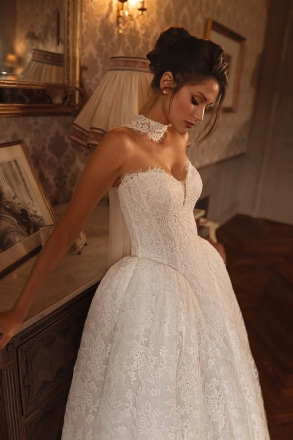 A woman in a wedding dress standing in an elegant room with vintage wallpaper, a framed black and white photo, and a table with a table lamp and mirror, looking down pensively.