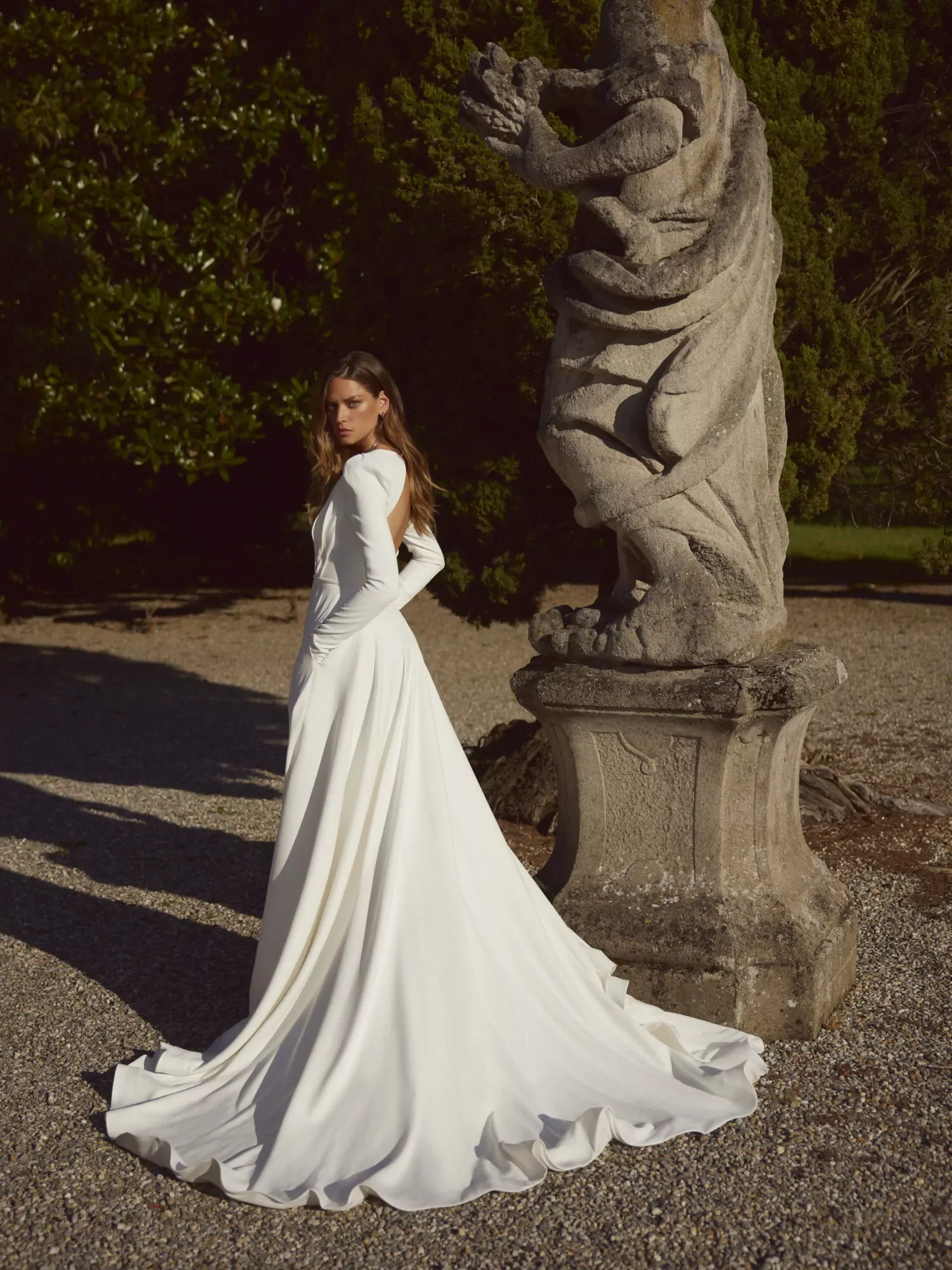 A woman in a long white dress with a train standing outdoors near a stone sculpture, with green trees in the background.