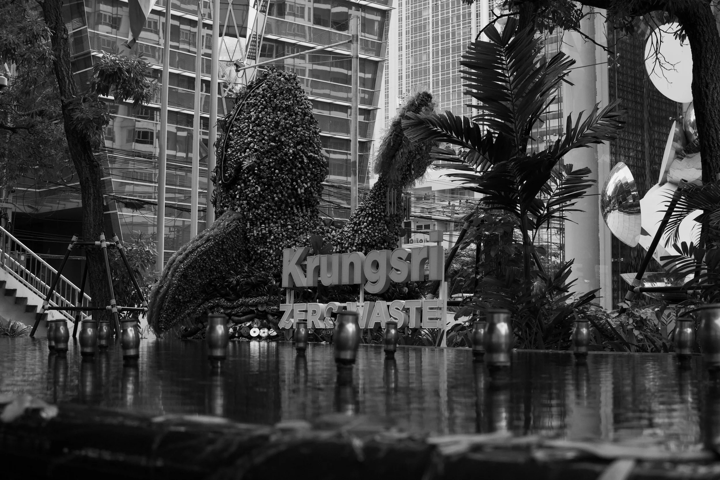 A large dinosaur sculpture made of trash and discarded objects, with the words 'Kungsan Zero Waste' nearby, surrounded by trees and modern city buildings in the background.