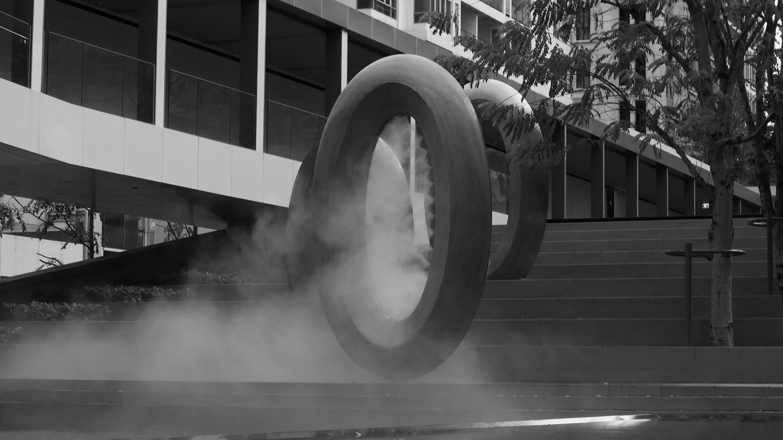 A large modern fountain with two circular rings and a central sculptural element emitting steam, located in an urban setting with glass buildings and trees around.