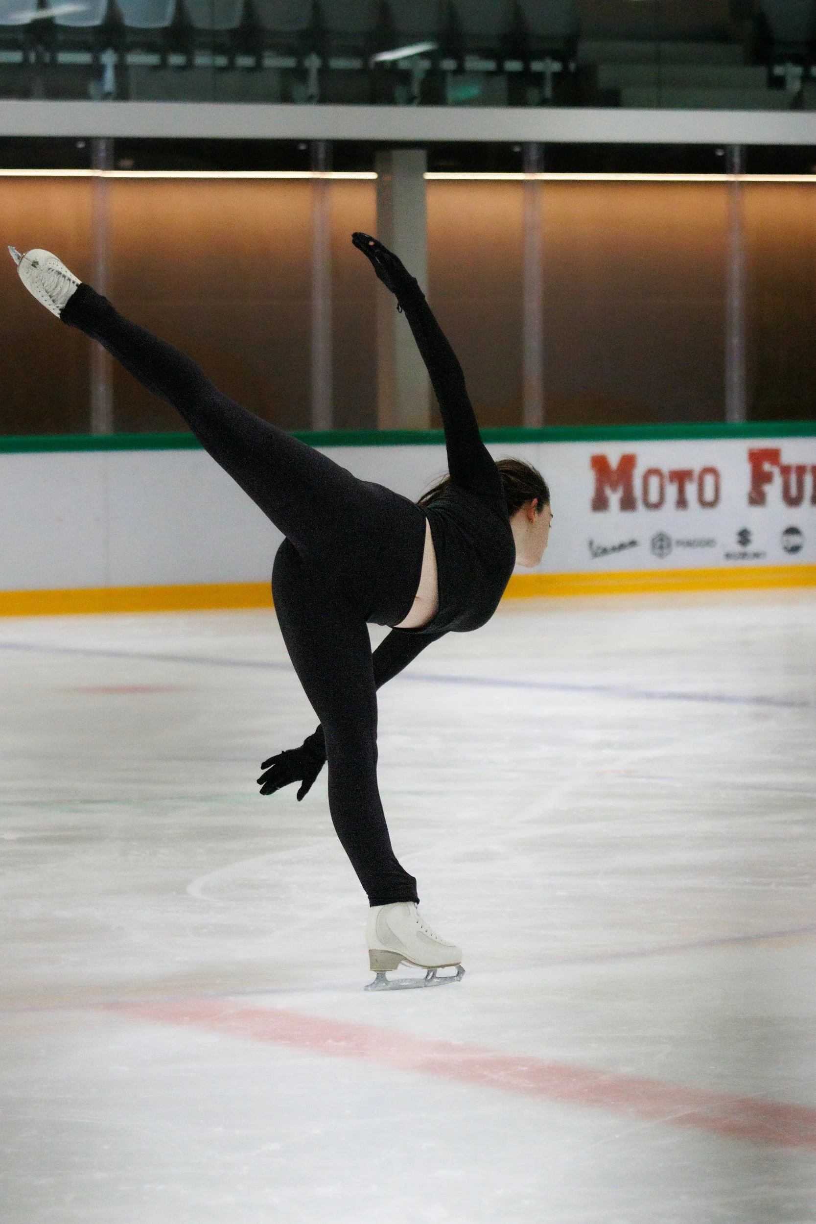 A female figure skater is performing a move on ice, standing on one leg with her other leg extended behind her and her arms raised. She is dressed in a black outfit with white figure skates.