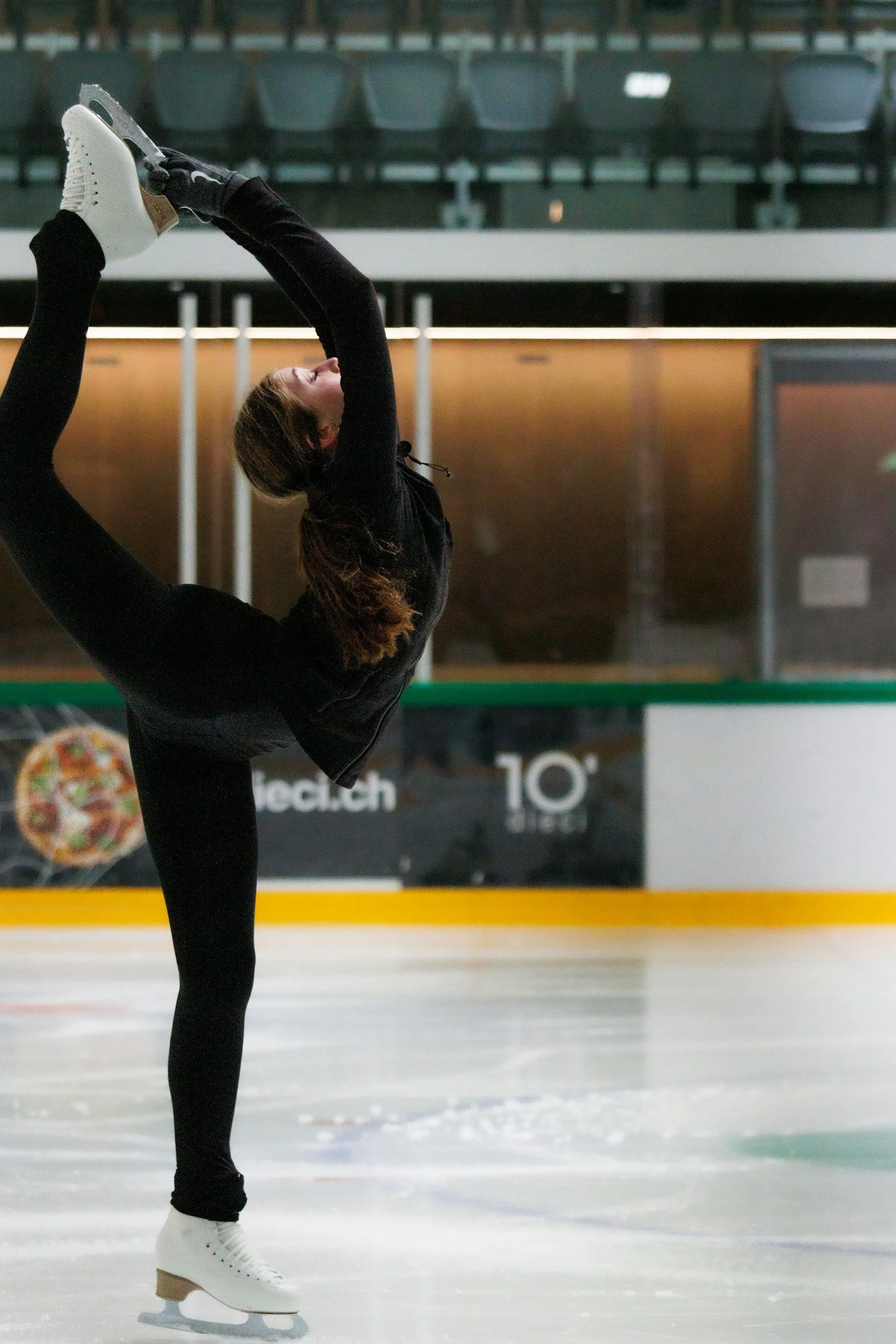 A female figure skater performing a move on the ice rink, with her leg extended above her head while balancing on one skate.