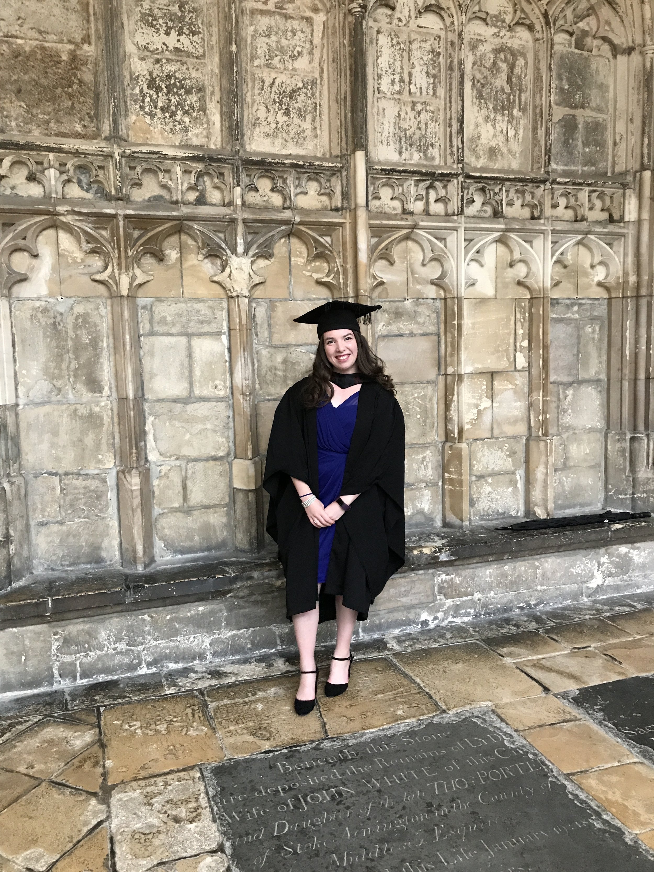 Young woman in graduation gown and cap standing in front of an old stone wall and engraved stone floor.
