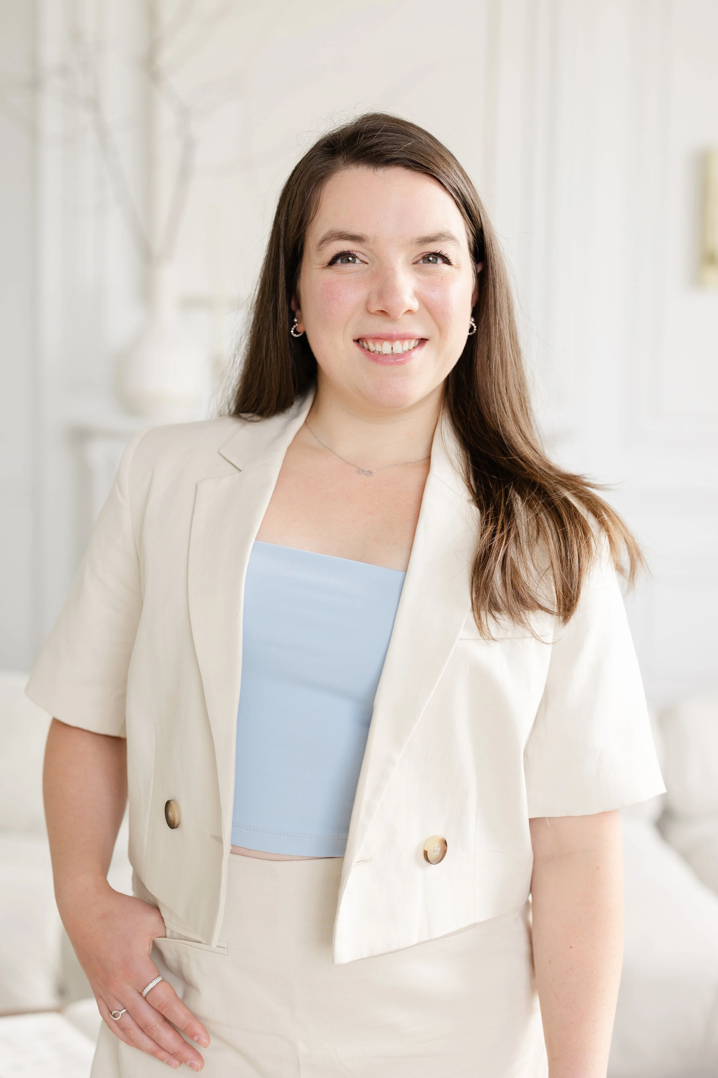 A woman with long brown hair, smiling, wearing a white blazer and a light blue top, standing in a bright, white room.