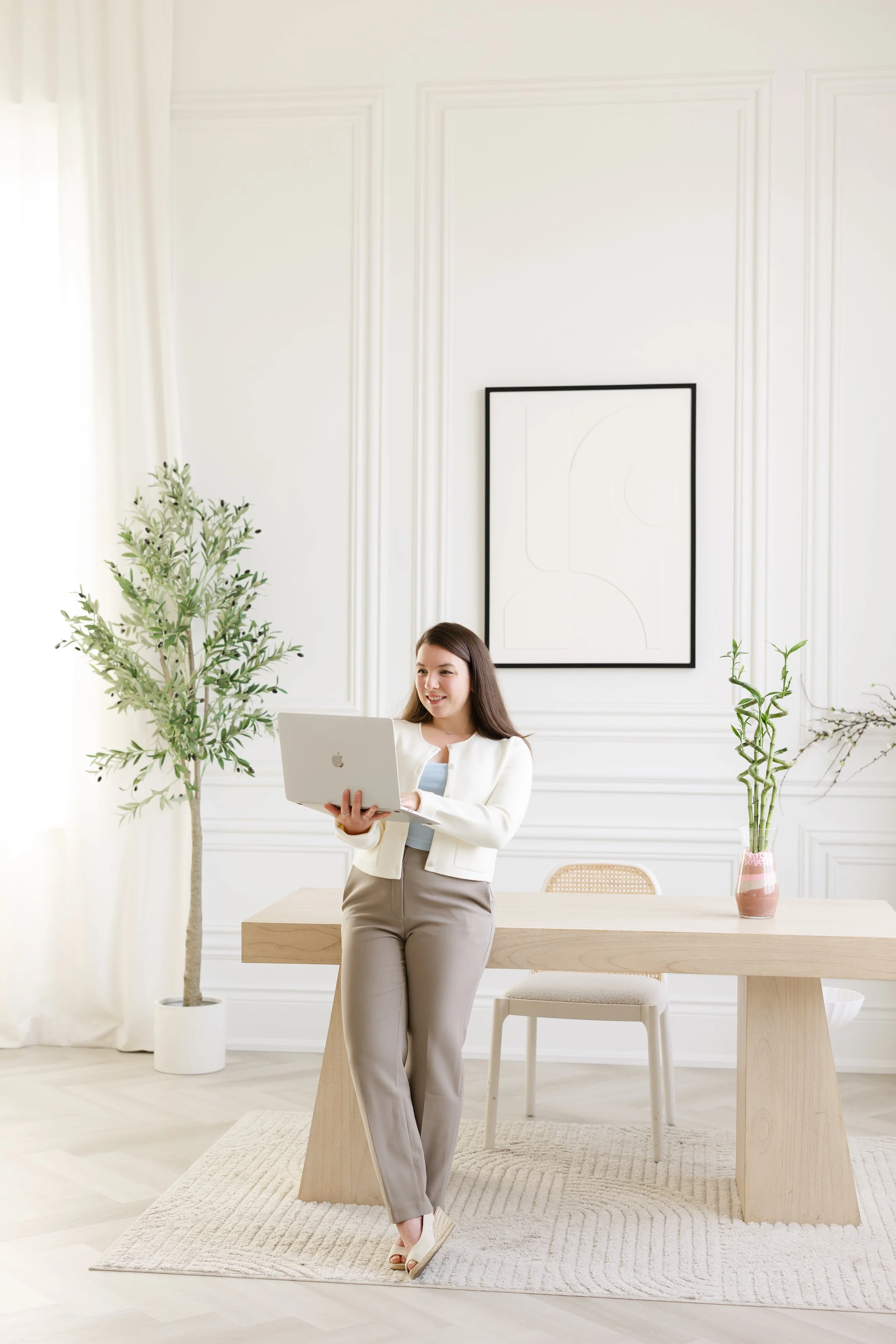 A woman in beige pants and a white blazer stands in a bright, modern, minimalistic room holding a laptop. The room features white walls, large leafy potted plants, and a wooden table with a pink vase, with natural light coming through a curtain.