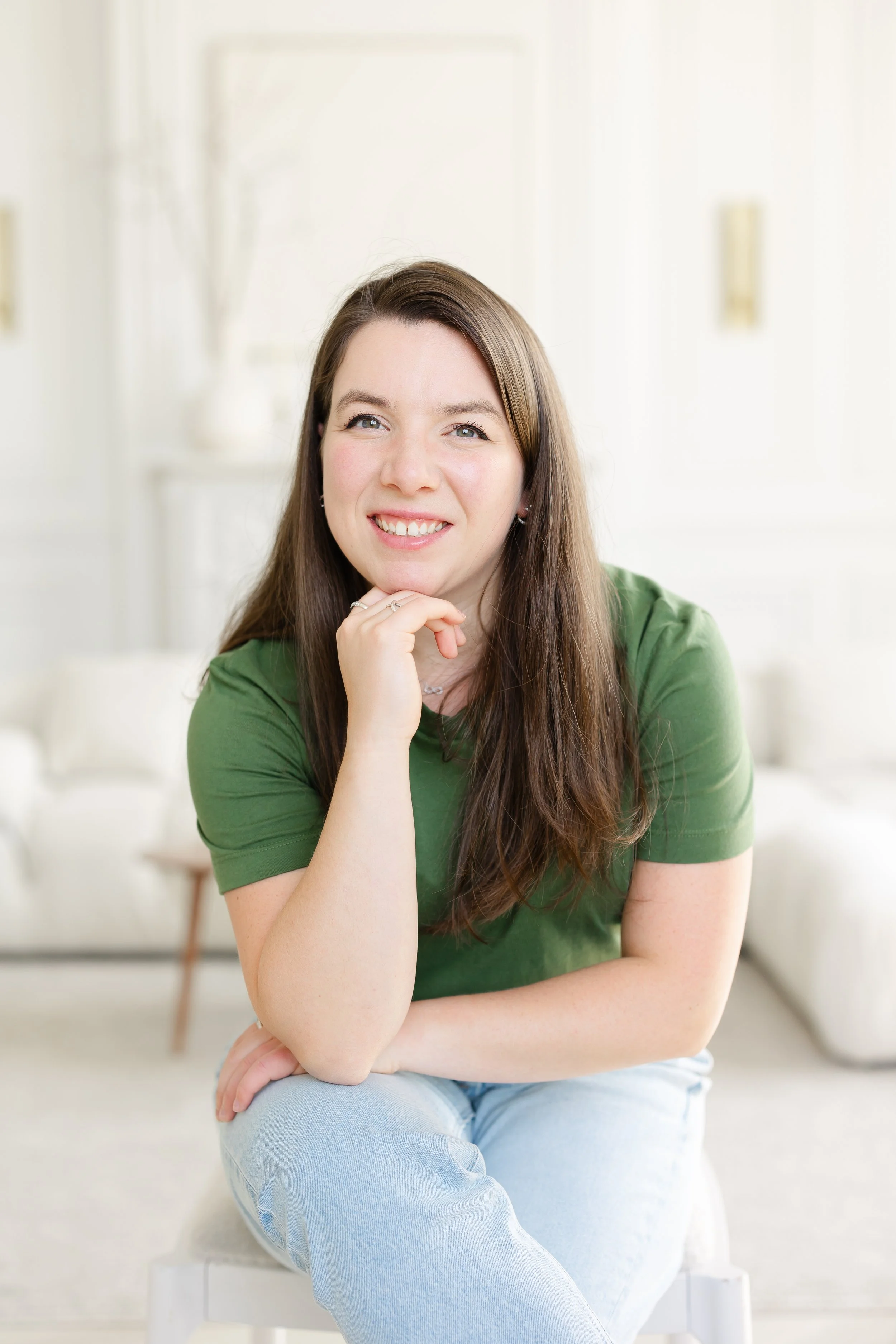 A young woman with long brown hair, wearing a green t-shirt and light blue jeans, sitting on a white chair in a bright, minimalistic room, smiling with her hand resting on her chin.