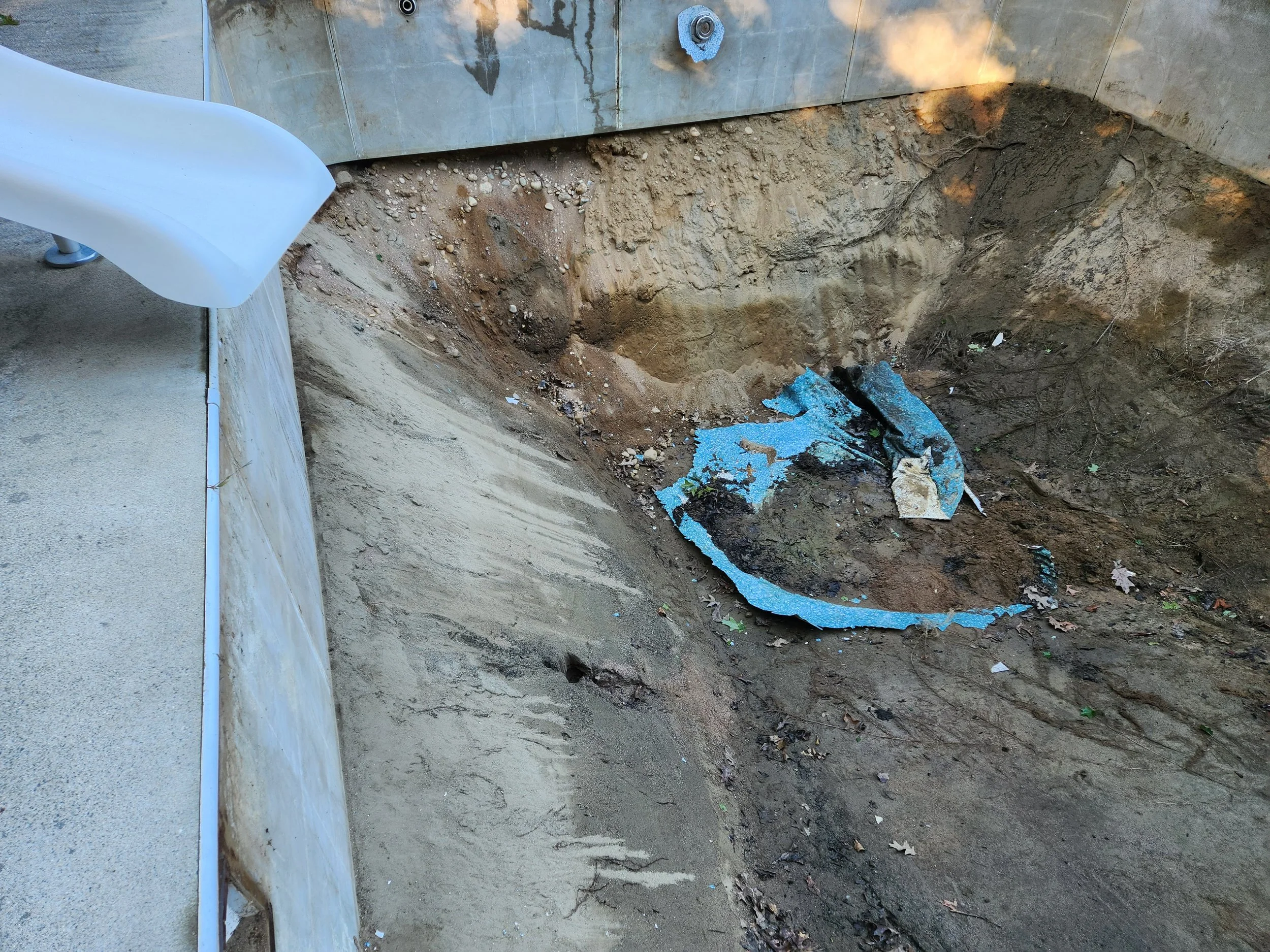 A large hole has been dug in the ground next to a building wall, with dirt and rocks around the sides and a piece of damaged blue foam insulation at the bottom. There are some scattered leaves and small debris inside the hole.