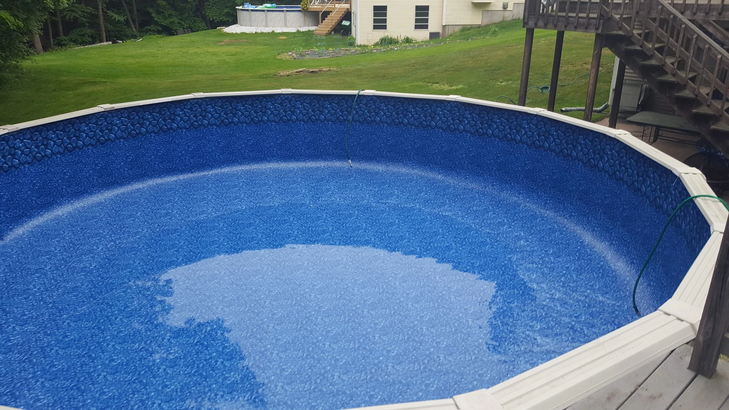 A blue above-ground swimming pool filled with water, situated on a wooden deck in a backyard, with a grassy area and a house in the background.