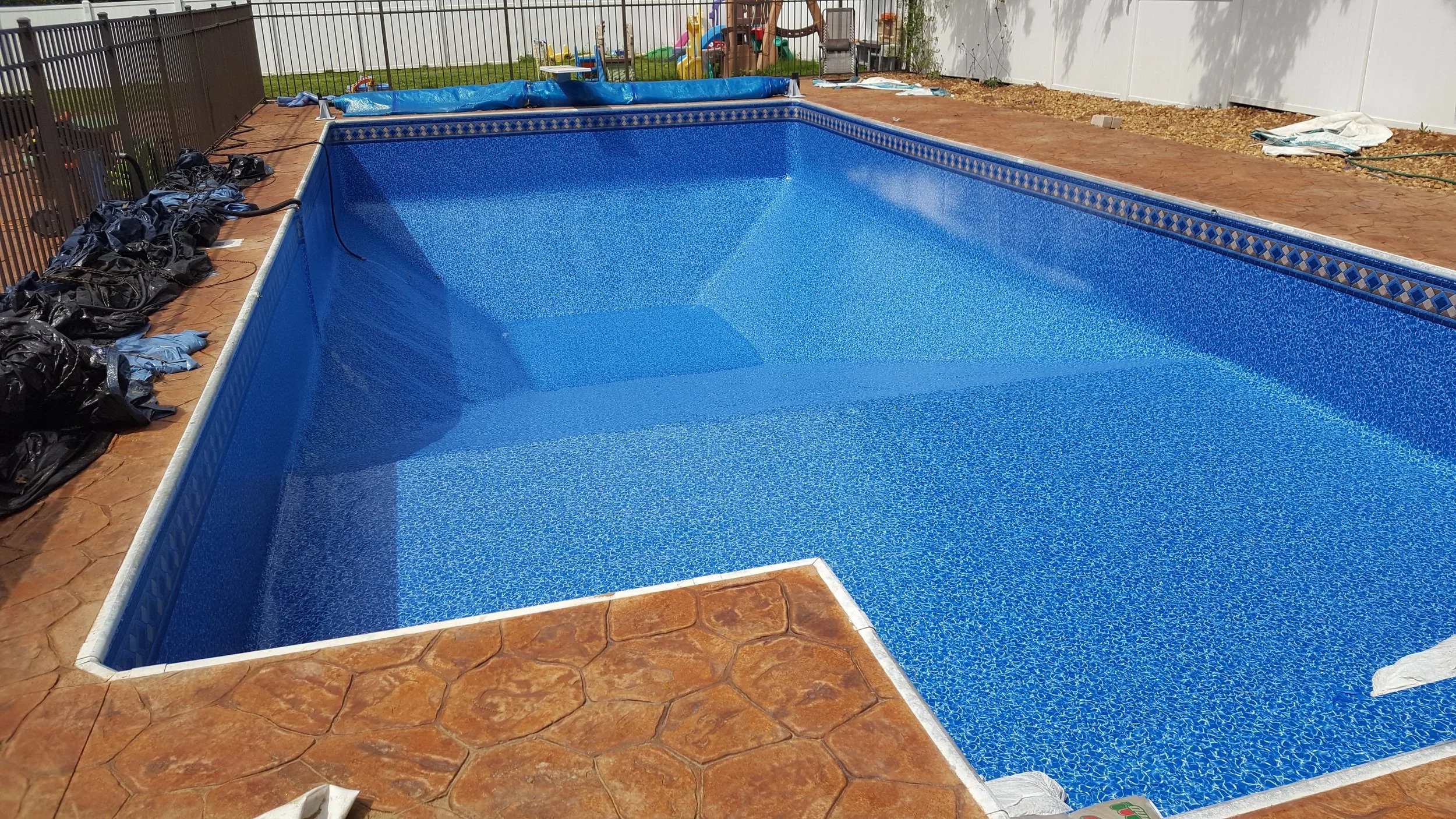 A rectangular in-ground swimming pool with blue mosaic tiles, surrounded by unfinished red brick paving and pool accessories, in a backyard enclosed by a white privacy fence.
