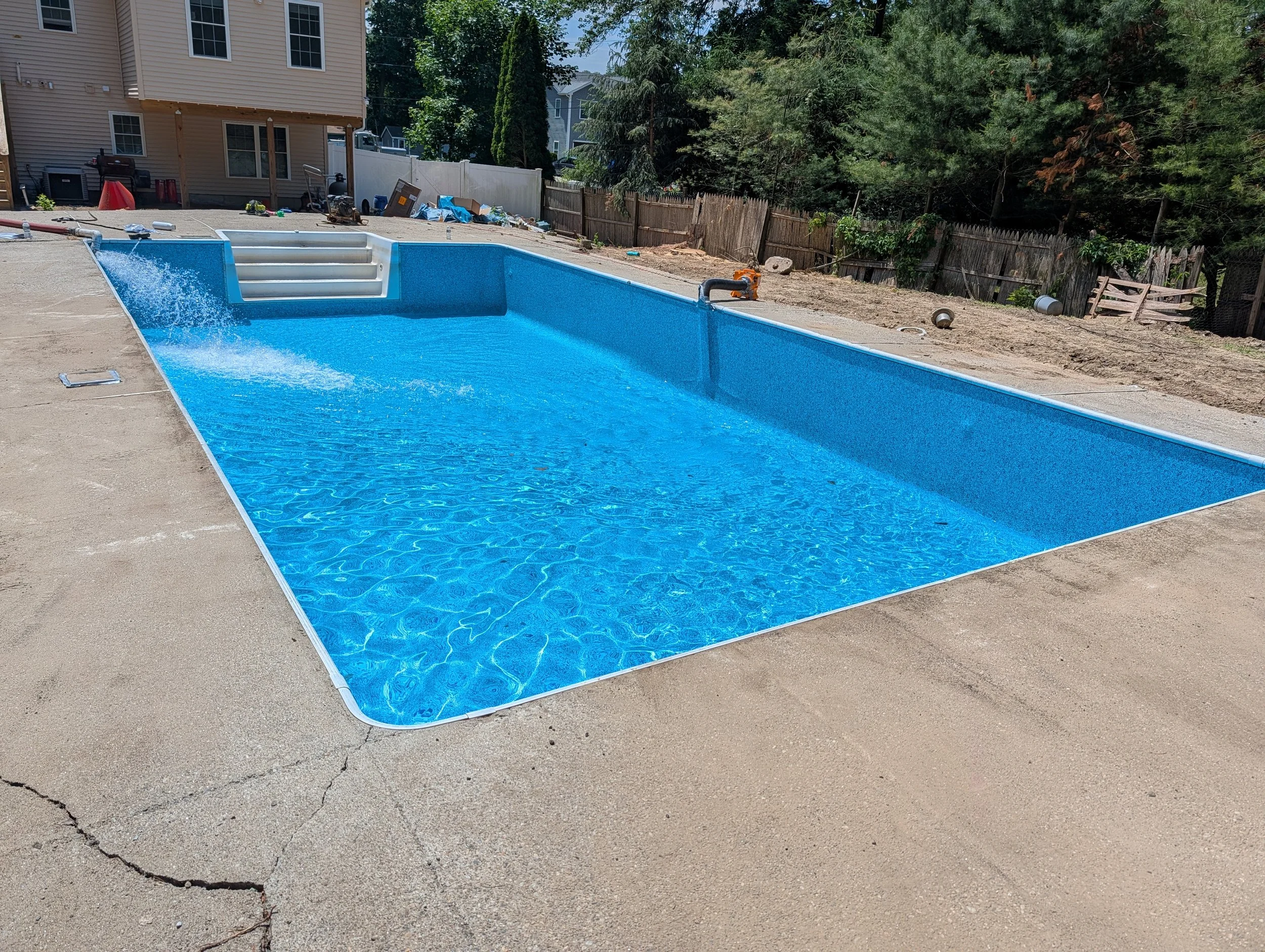 A rectangular swimming pool with blue interior and white steps under construction in a backyard, surrounded by unfinished concrete surface and debris, with a house and trees in the background.