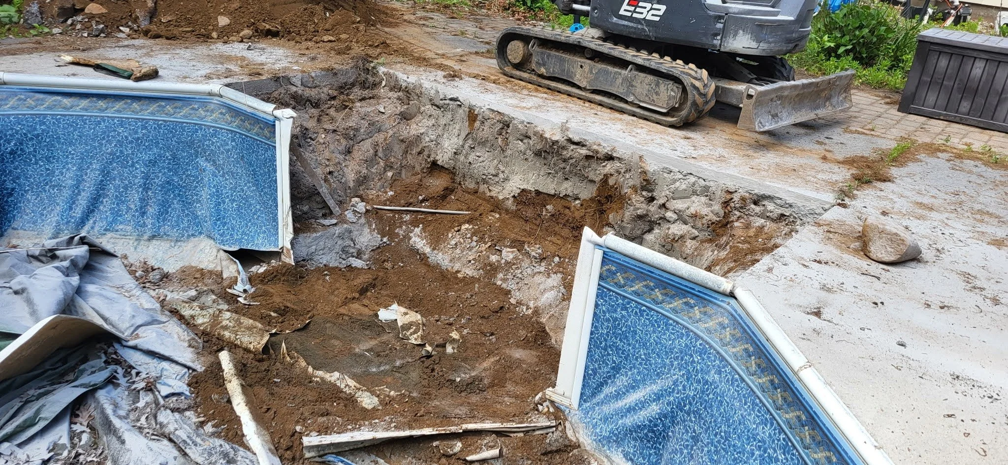 A construction site showing a destroyed swimming pool with blue patterned tiles. There is a large hole in the ground, dirt, and construction debris. An excavator is parked nearby on a partially dismantled concrete surface.