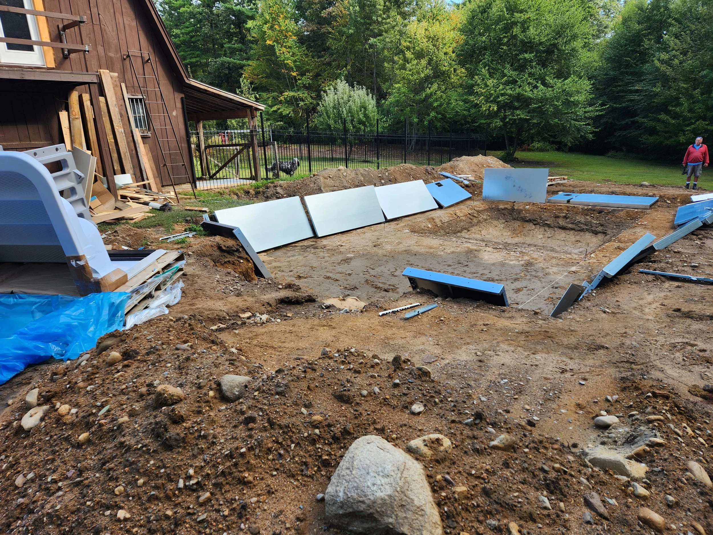 A backyard under construction with exposed dirt and scattered construction materials, including large white panels, scattered around a dug-out area. A man in a red jacket and shorts is walking on the right side, while a dog stands near a fence. There
