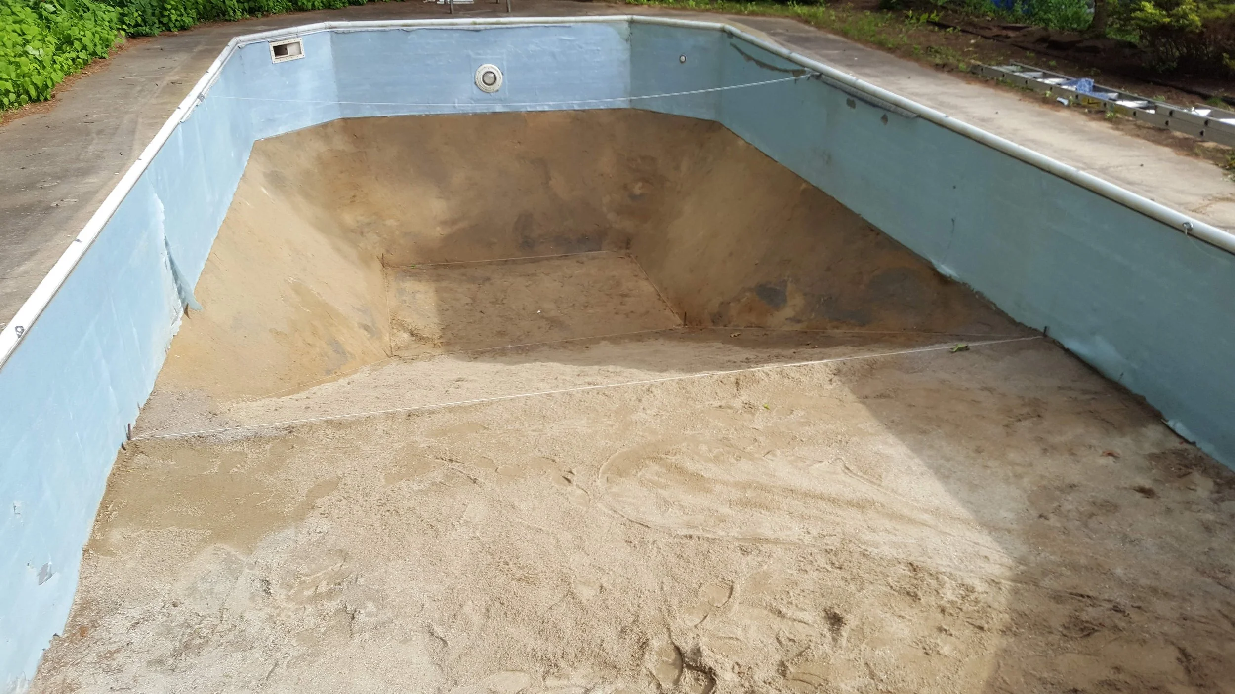 An empty swimming pool undergoing construction or renovation, with sand at the bottom and sides, and some construction tools and materials visible on the ground nearby.
