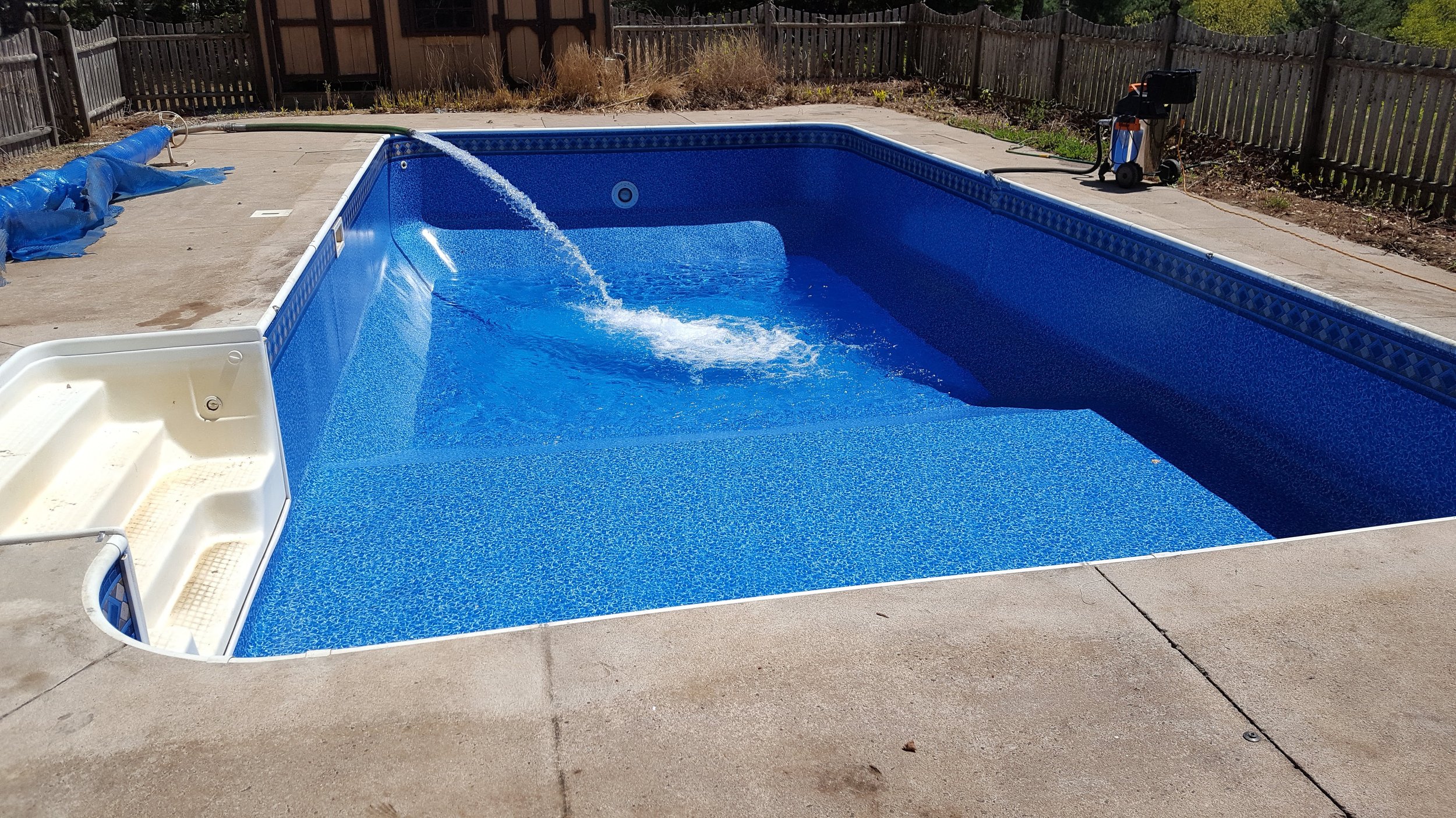 Empty backyard swimming pool with blue interior, water flow from a hose, a pool vacuum on the deck, and a wooden fence surrounding the yard.