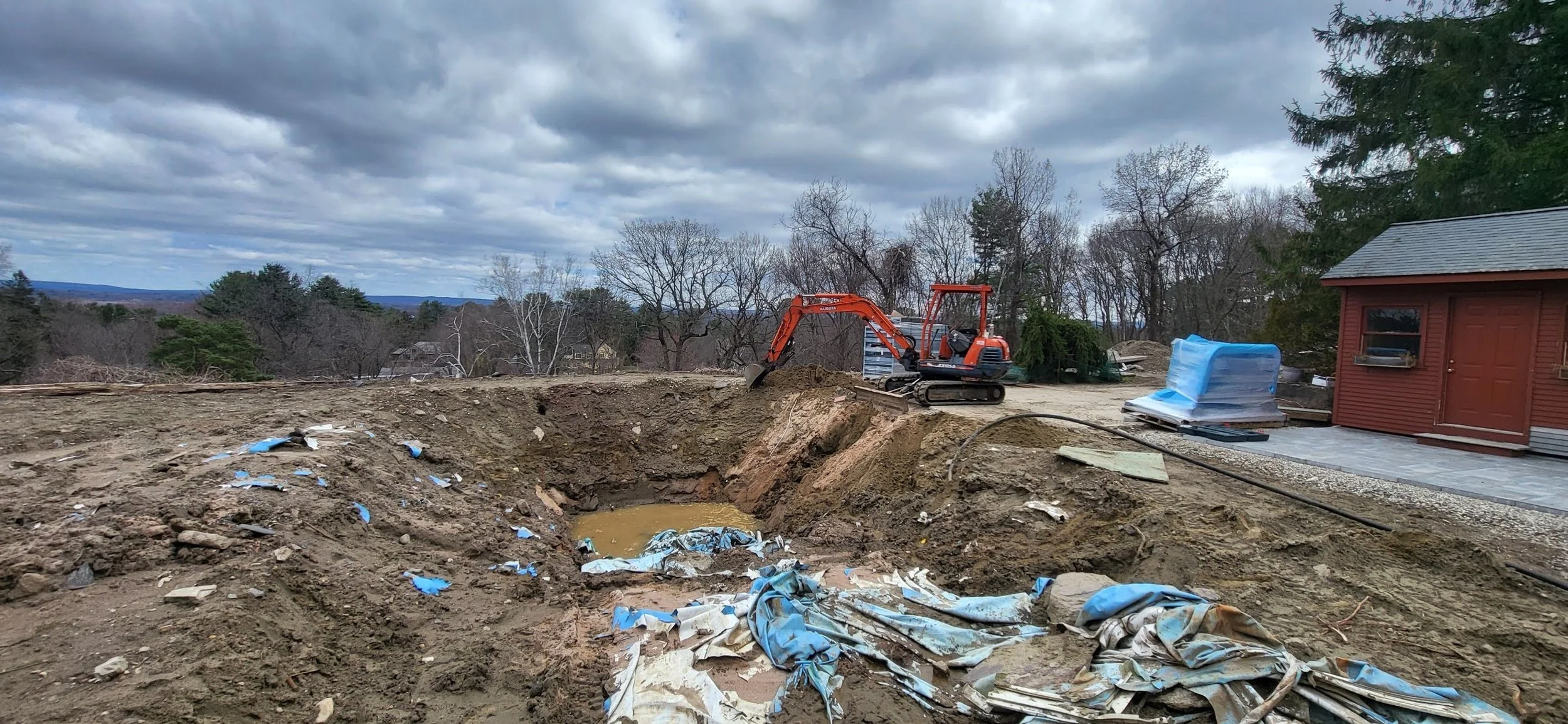 An outdoor construction site with a small orange excavator digging a trench, surrounded by dirt, construction debris, and blue plastic sheeting. There is a red building and trees in the background under a cloudy sky.